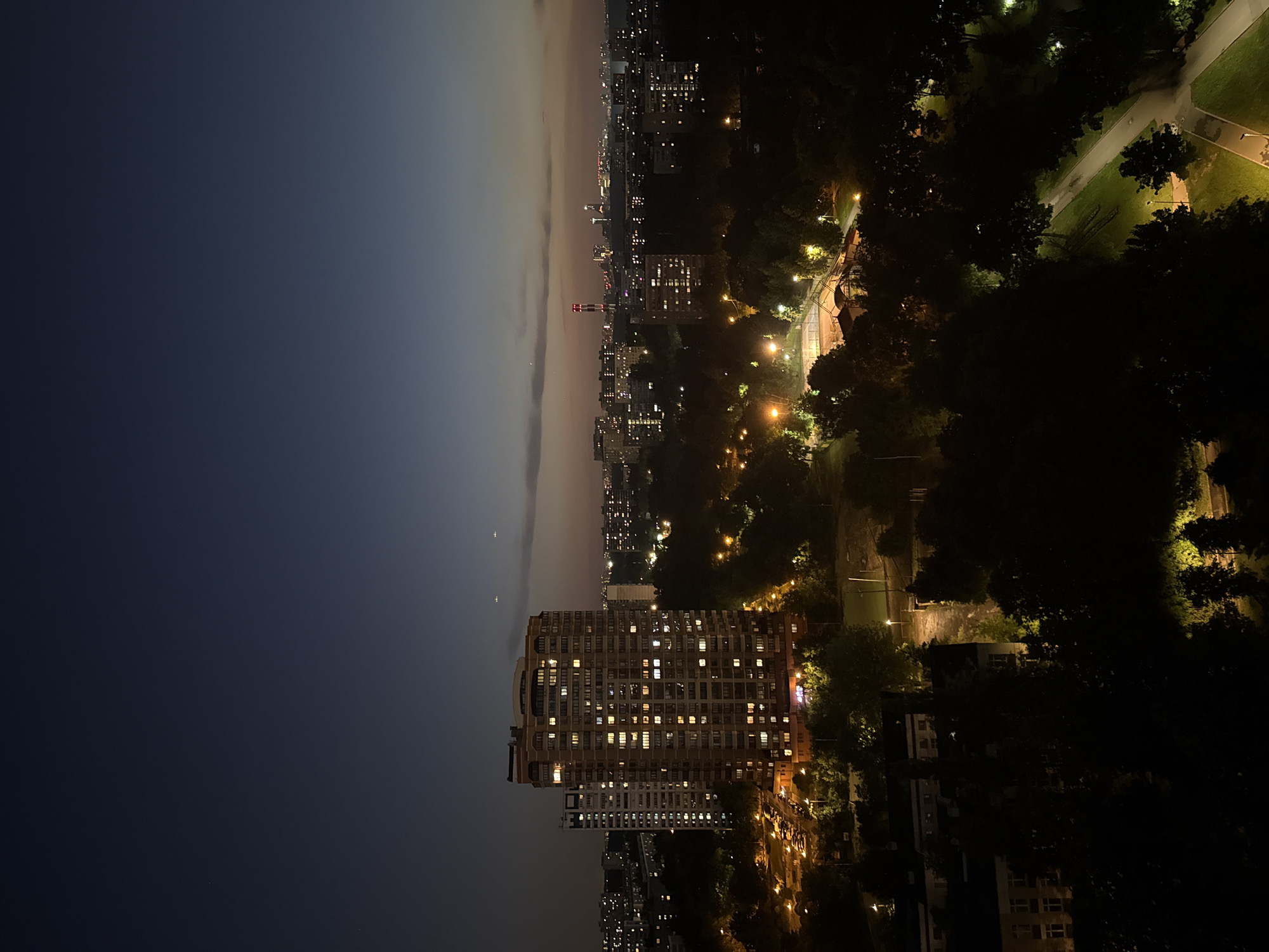 An aerial view captures a nighttime city scene, likely in Moscow, Russia, bathed in the fading twilight. The sky transitions from a deep, dark blue at the zenith to a lighter, dusky hue near the horizon, with a few faint celestial bodies visible. Prominent in the foreground is a tall, multi-story apartment building with numerous windows illuminated by interior lights. Adjacent to it stands another, lighter-colored apartment building. The middle ground features a dense canopy of trees, some illuminated by streetlights, interspersed with buildings and the faint glow of distant city lights. A sports court, possibly a tennis court, with bright artificial lighting, is visible amidst the greenery. In the far distance, the city skyline is dotted with numerous lights, and a red-and-white striped tower with illuminated white bands stands out. The overall impression is one of a residential area within a larger urban sprawl at dusk. There are no visible people or animals. The scene is static, depicting a quiet, urban night.