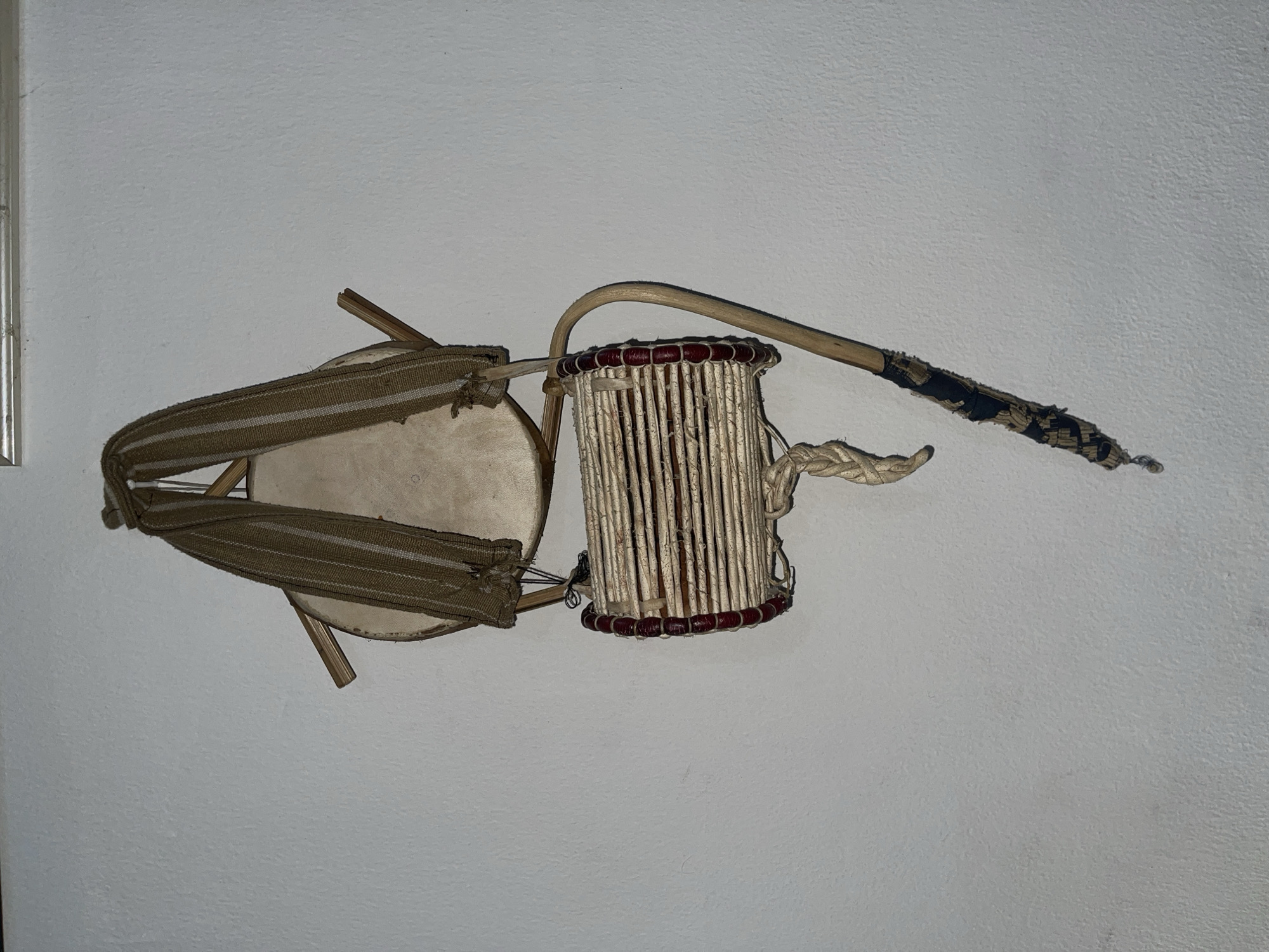A still life photograph depicts a talking drum, which is a type of West African drum, resting against a plain white wall. The drum consists of two drumheads, each circular, and interconnected by a series of cords and a central, cylindrical resonator. The left drumhead has a beige, leather-like appearance with a broad, striped strap running over its surface and secured to the wooden structure underneath. The right drumhead is comprised of vertical, light-colored slats held together by a red-beaded rim. A curved, wooden stick with a frayed, dark-colored covering on the handle end is also present, positioned to the right of the drum. A rope-like element is tied near the base of the drum, adding to the texture of the composition. The photograph is taken from a high-angle perspective, casting slight shadows on the wall, which suggests the lighting is from above. There is no indication of the location being in Ado, Nigeria.