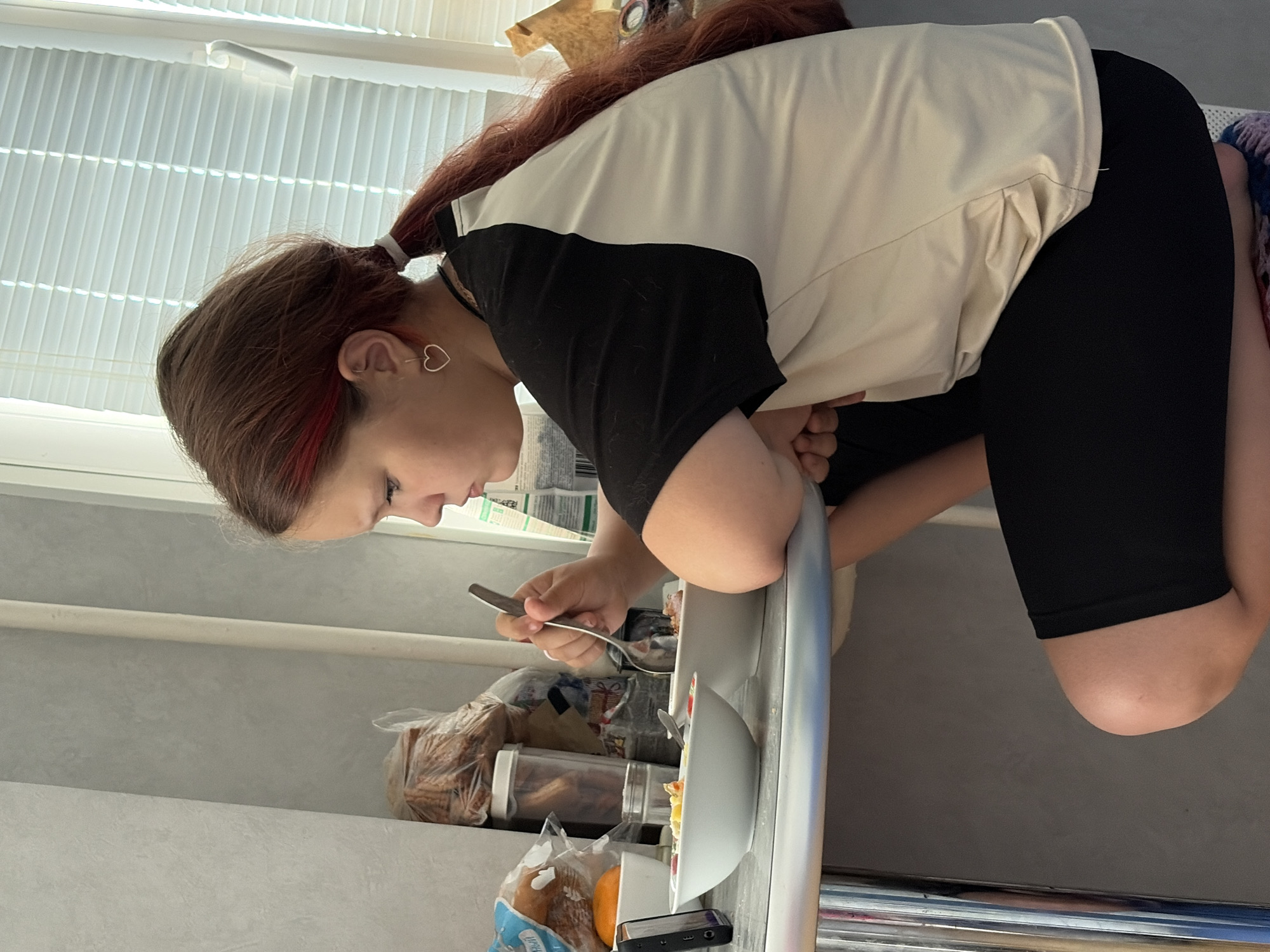 A young person with long, reddish-brown hair tied in a ponytail is shown leaning over a light gray countertop, eating from a bowl with a spoon. A distinctive red streak is visible in her hair. She wears a heart-shaped earring in her left ear and is dressed in a two-toned shirt (black sleeves and upper back, light beige body) and black shorts or leggings. Her posture suggests she is kneeling or sitting low beside the counter.
On the countertop, two bowls of food are visible, one white and one light gray, each with a spoon. Behind these, various items are arranged, including several plastic bags of baked goods (likely bread), clear food containers, and a black smartphone. Text on one of the plastic bags reads "ЛУКОШКО," a Russian word meaning "basket."
The scene is brightly illuminated by natural light entering through white pleated window blinds in the background, indicating it is daytime. A light-colored horizontal bar spans the wall behind the counter. A glimpse of a colorful, possibly knitted, fabric or blanket can be seen near the person's right leg. The setting appears to be an indoor space, likely a kitchen or dining area, in Chaikovsky, Russia, as suggested by the visible text and provided location context. The overall impression is one of a quiet mealtime.
