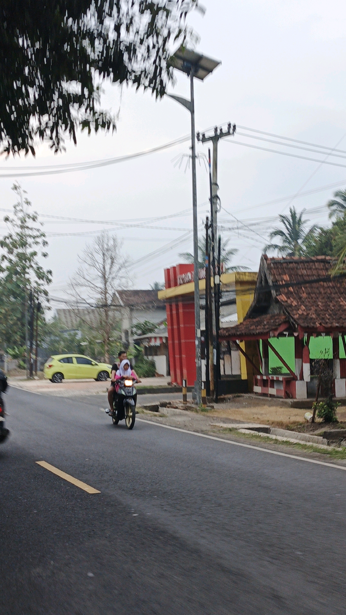 The image depicts a street scene in Cibenda, Indonesia. In the foreground, a dark asphalt road with a yellow dashed line runs diagonally from the lower left to the upper right. A motorcycle with two occupants, a male driver and a female passenger wearing a pink hijab, travels away from the viewer on the right side of the road. The male appears to be wearing a dark shirt and the female a lighter colored garment. Behind them, to the left of the road, a bright yellow car is parked.
Further back, several buildings are visible. On the left, a building with a red facade and yellow trim stands behind the yellow car. To the right of this, a smaller structure with a tiled roof and green window frames is situated closer to the road. A large utility pole with multiple wires stretches across the frame behind the motorcycle and buildings. Trees with green foliage are present on the left and right sides of the image, with some bare branches visible in the center. The sky is overcast and light grey. The overall scene appears to be a typical street in a populated area.