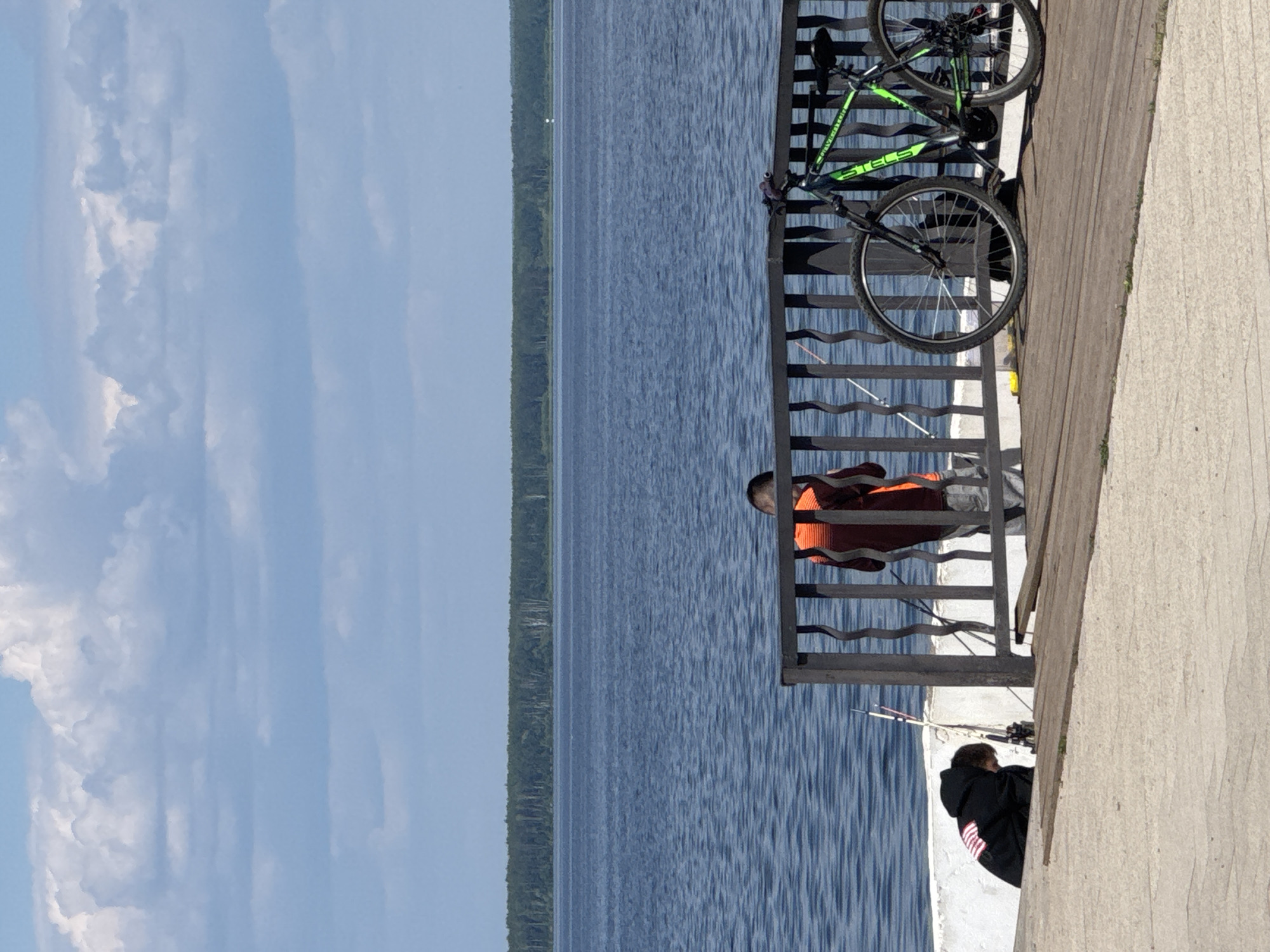 The image depicts a scene near the Kama River in Chaikovsky, Russia, on a clear day. In the foreground, a wooden pier extends towards the river. Two individuals are visible. One, wearing a black hooded sweatshirt with an American flag emblem on the back, is bent over near a white retaining wall, appearing to be fishing. Several fishing rods are propped up nearby. Further along the pier, another individual, seen from the back, is wearing an orange life vest over a dark top and light-colored pants, also seemingly engaged in fishing. A black and green bicycle is parked on the pier next to a metal railing. The background features the calm, blue waters of the Kama River, with a distant treeline along the horizon under a sky filled with scattered white clouds.