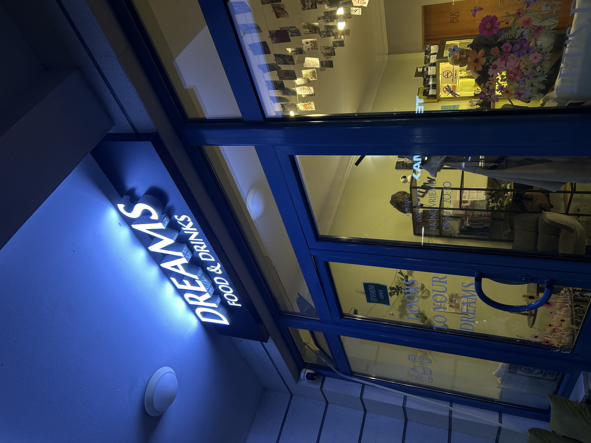 The image depicts the entrance to "Dreams Food & Drinks," a café bar in Budva, Montenegro. The entrance features a blue frame around a glass door and windows. Above the entrance, a neon sign reads "Dreams Food & Drinks." The glass reflects the interior, showing a shelf with books and other objects, a flower arrangement, and hanging photographs or cards. A sign saying "OTVORENO OPEN" is on the glass door. The sign's text reads in Montenegrin and English.