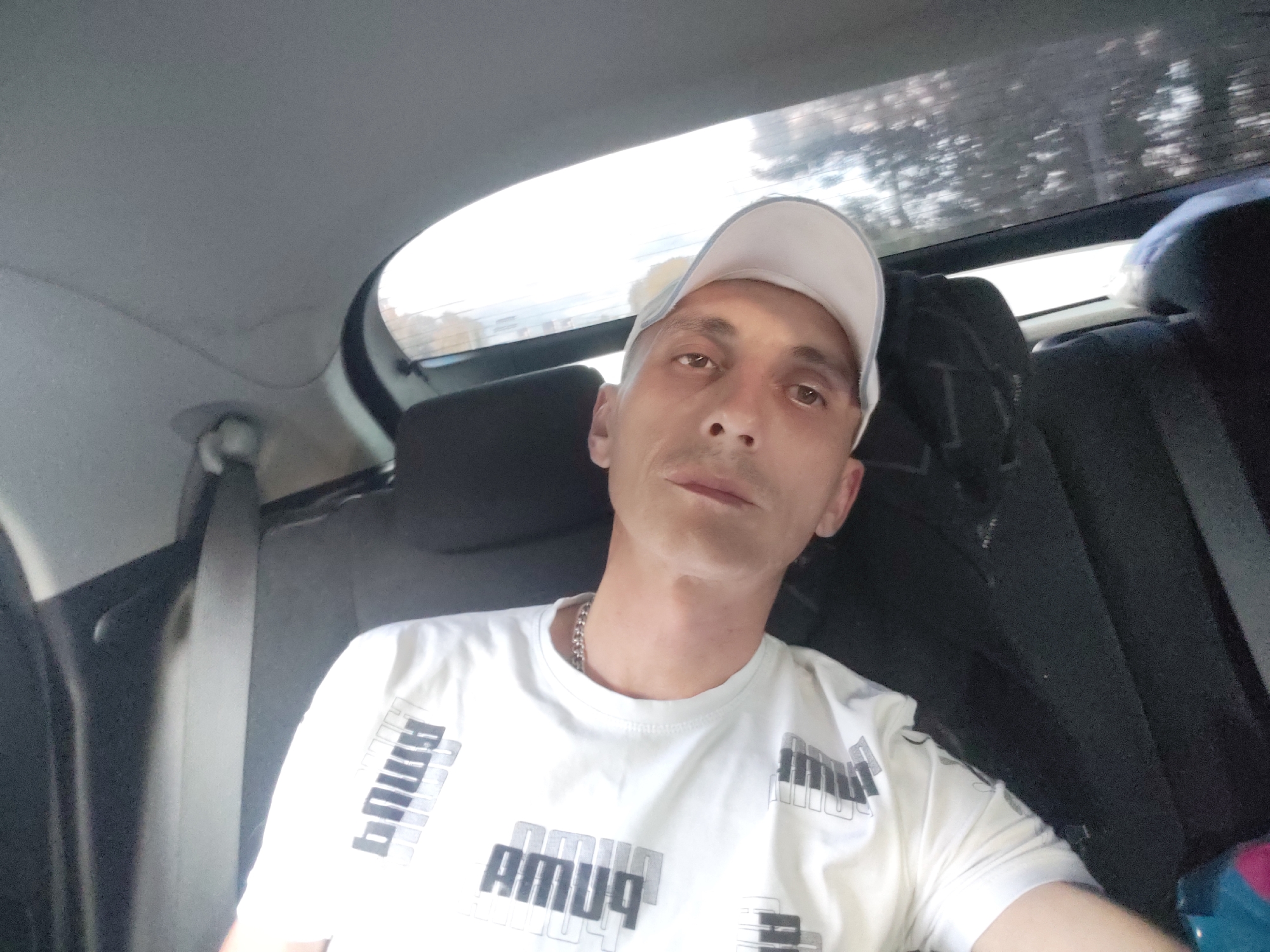 In this close-up, self-taken photograph, a Caucasian man with short, light-colored hair and a light-colored baseball cap is seated in the back seat of a car. He is wearing a white t-shirt with a repeating black and white "PUMA" logo design. He has a silver chain around his neck. The background shows the interior of a car, including the headrests of the seats and a portion of the rear window, through which trees and a bright sky are visible. The image appears to have been taken during daylight. There is no specific visual information within the image to determine the exact location within the city of Bor, Russia.