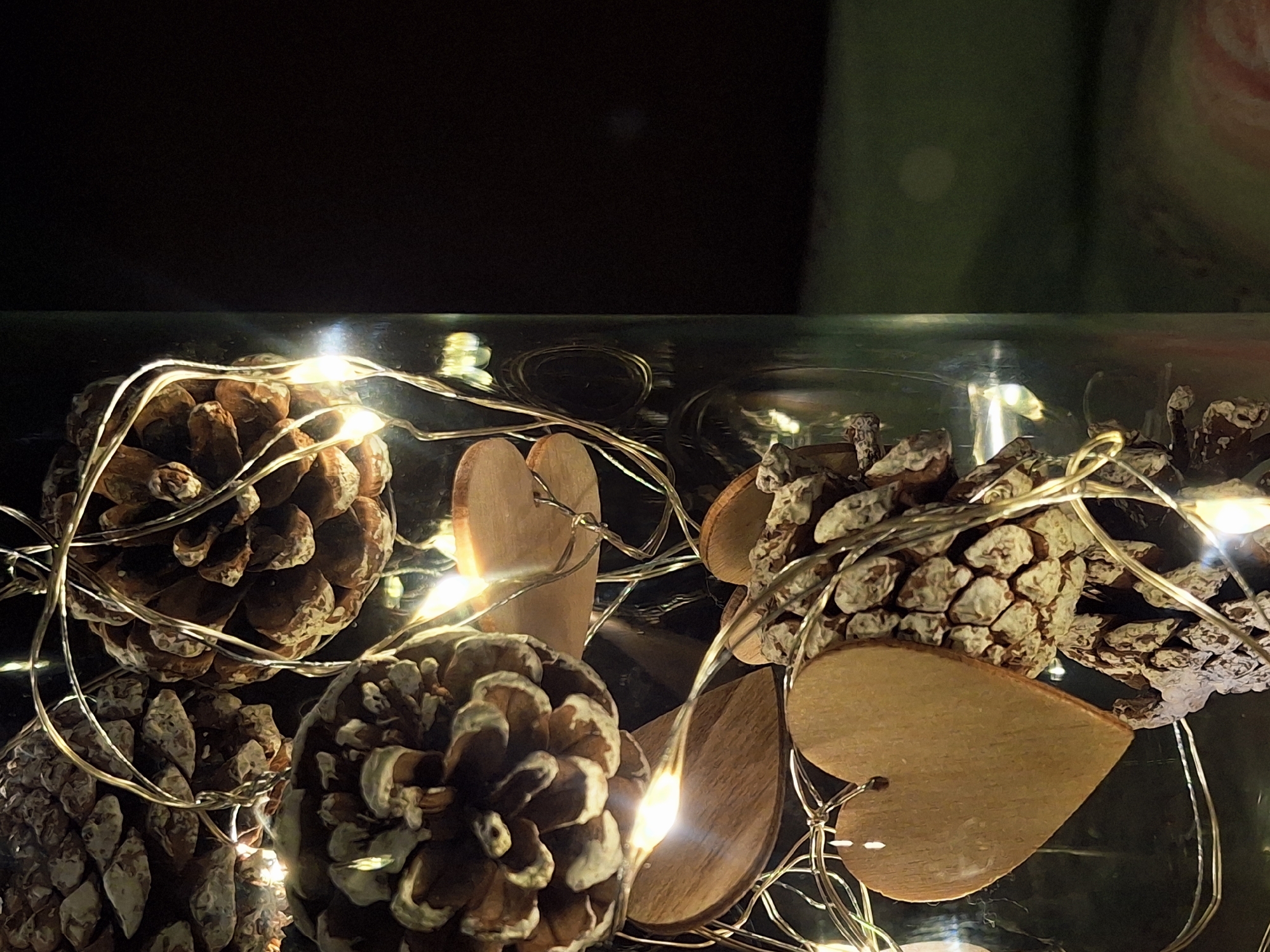 The image depicts a close-up view of a decorative arrangement inside a clear glass container. The arrangement consists of several pine cones, and wooden heart shapes. A string of small, warm-toned lights is intertwined among the objects, adding illumination and creating a festive or cozy ambiance. The background is dark, with a slight green tint visible at the top right.
There is no information in the image about the location in the city of Chaikovsky, Russia.