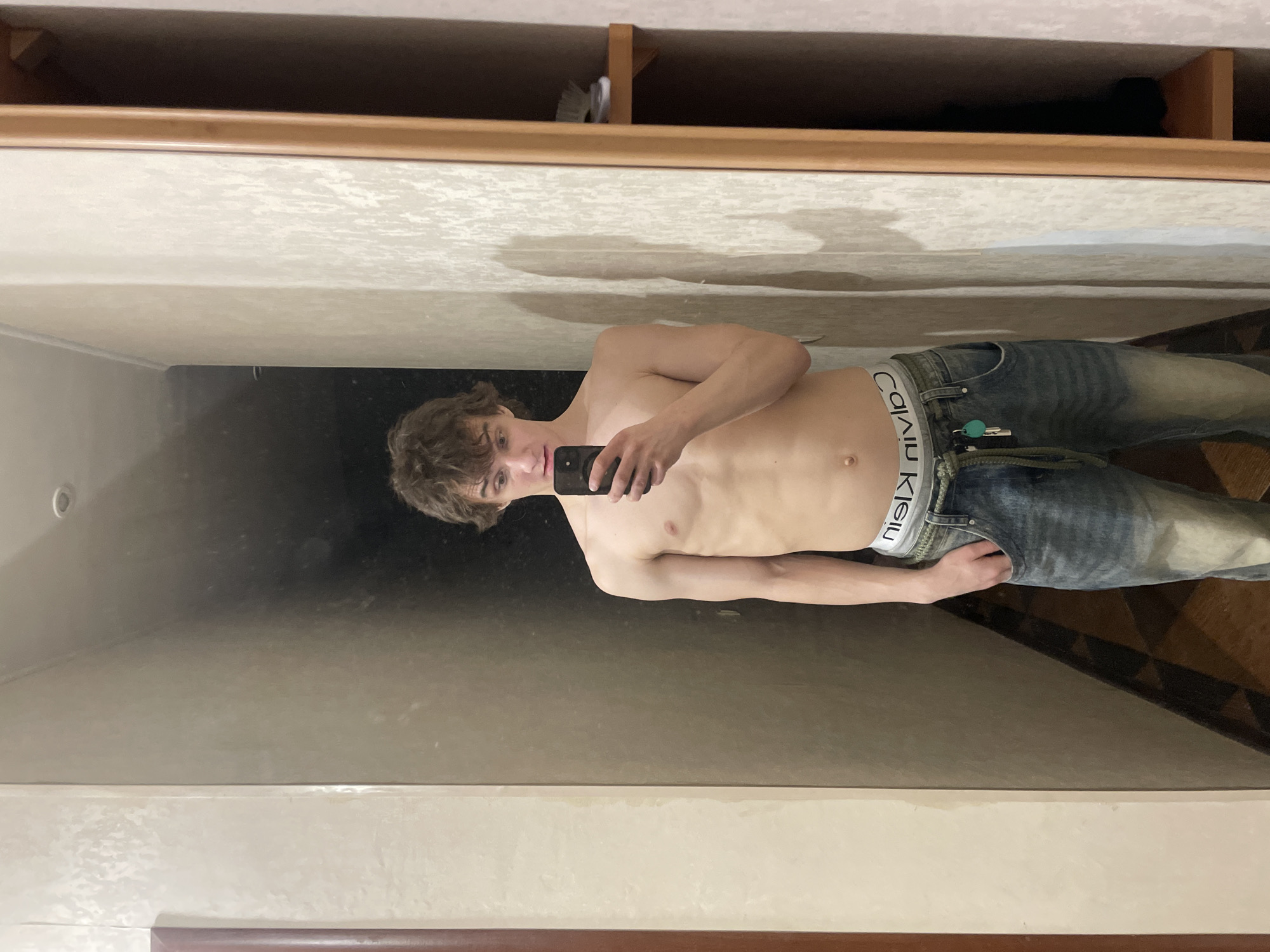 A young, shirtless male with curly brown hair is taking a mirror selfie. He is wearing blue jeans with a visible Calvin Klein waistband. The reflection reveals a bathroom setting with a vanity cabinet above the mirror and tile flooring partially visible in the lower right. The location cannot be determined as Krasnoyarsk, Russia, from the available visual information.