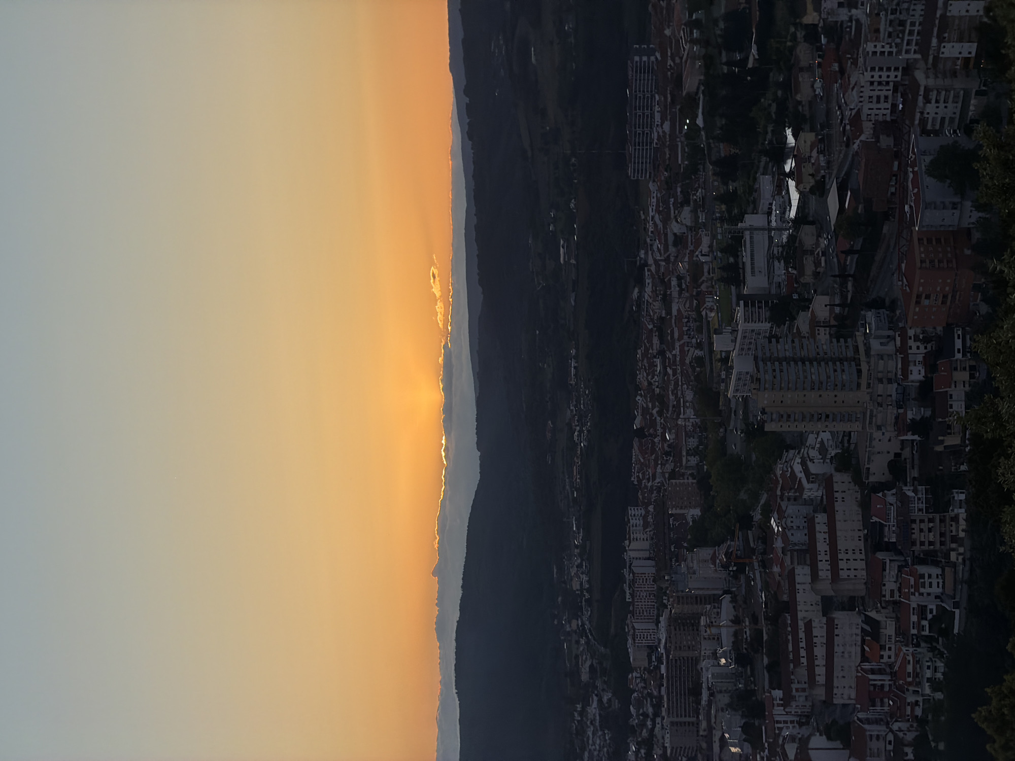 The image captures a cityscape at sunset, likely in Bragança, Portugal, given the presence of distinct architectural features common to the region. The composition is divided vertically: the left side depicts the sky and setting sun, with golden rays filtering through a mountain range or hill, while the right side shows the city below. The city consists of buildings of varying heights and architectural styles, densely packed together. The lack of visible human activity and the dim lighting on the cityside suggest either late evening or a deliberate shadow effect. The skyline is dominated by residential and commercial buildings, possibly with a mix of older and newer structures.