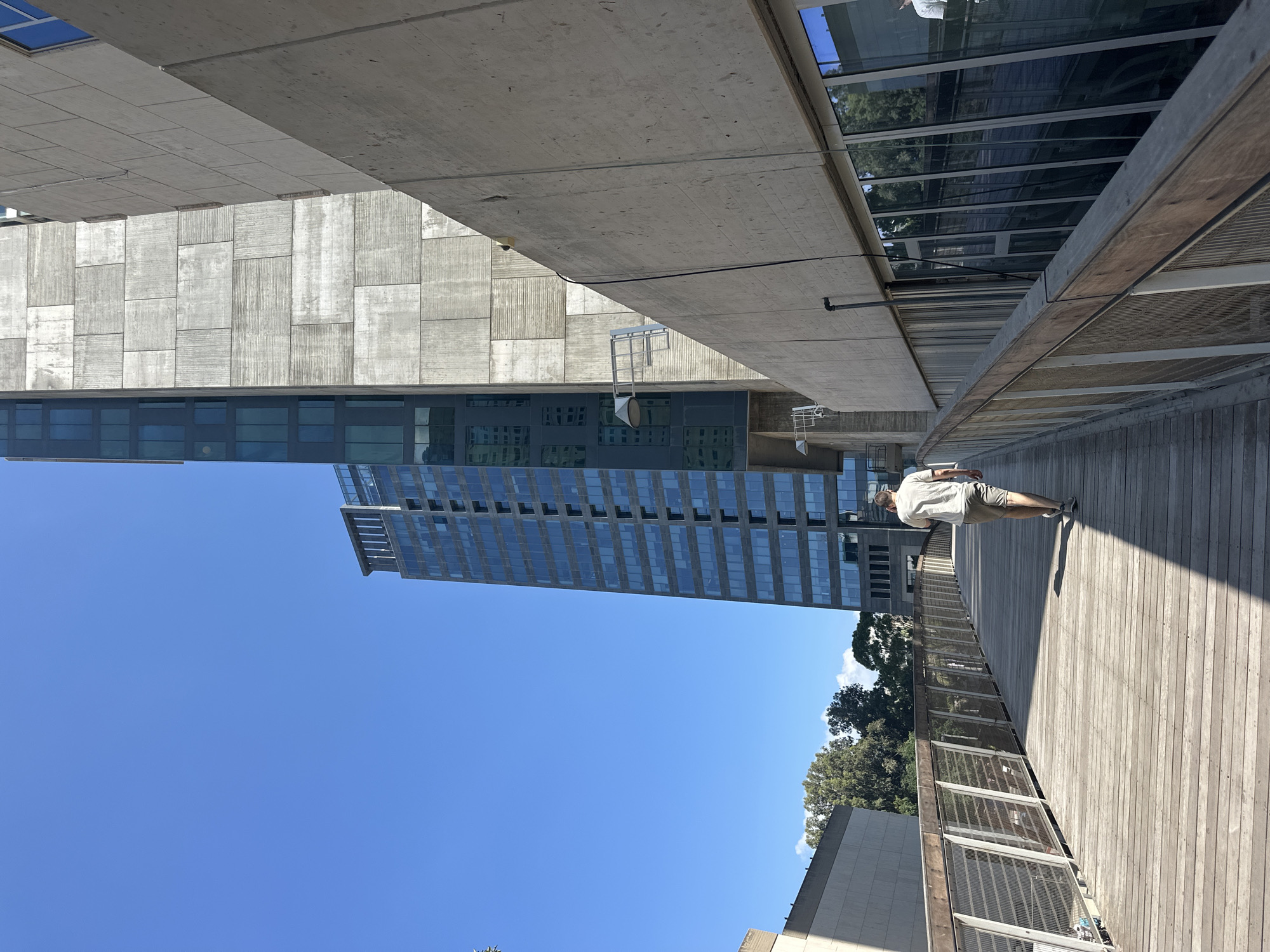 A person walks on a wooden pedestrian bridge in Tel Aviv, Israel. The bridge has metal railings and wooden planks. To the right of the bridge is a modern building with a concrete facade and large windows. Further in the background, another tall glass-faced skyscraper is visible, reflecting the clear blue sky. Trees are present on the left side of the scene. The person is wearing a light-colored t-shirt and shorts. The sun casts shadows across the bridge, indicating it is daytime.