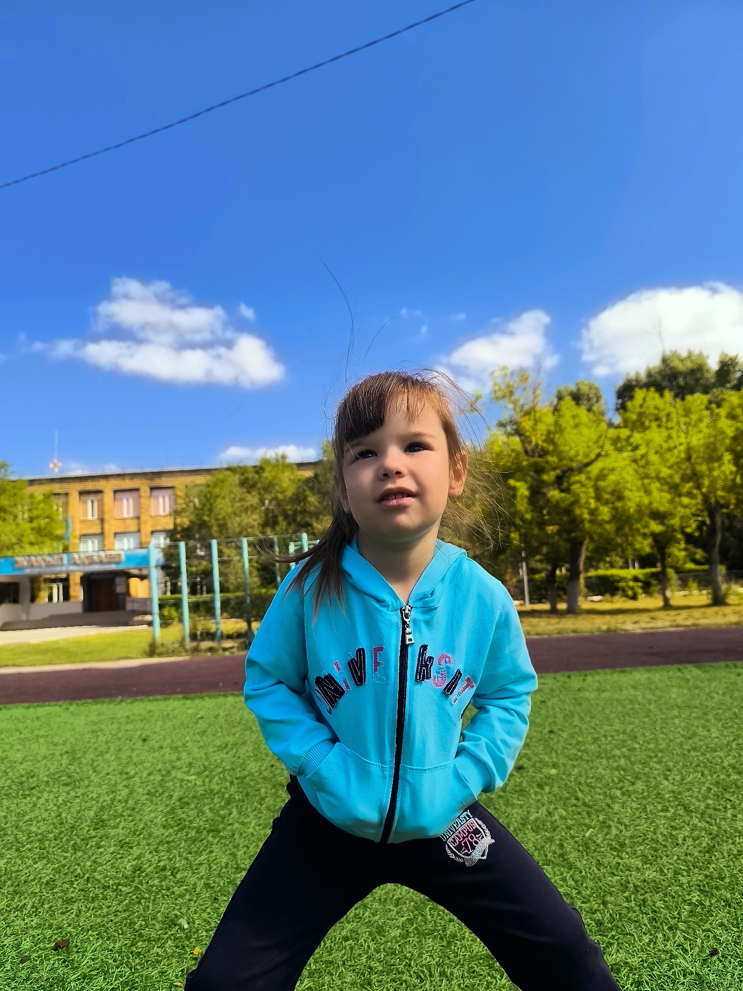 A young girl stands on a bright green artificial turf field, her hands tucked into the pockets of a light blue zip-up hoodie. Her dark hair is pulled back in a ponytail, and she looks up and to the right with a curious expression. The hoodie has pink and black lettering on it, and her black pants have a crest-like emblem on the left thigh.
Behind the girl, a building with a yellowish facade and multiple windows is visible. A blue sign with white lettering hangs above the entrance. To the right of the building, several green trees are in bloom, their leaves a vibrant green. The sky above is a clear, bright blue with a few white clouds and a thin, dark line that appears to be a wire stretched across it. The scene is outdoors, likely in a park or schoolyard, given the artificial turf and the presence of the building and trees. The setting suggests a sunny day in a city.