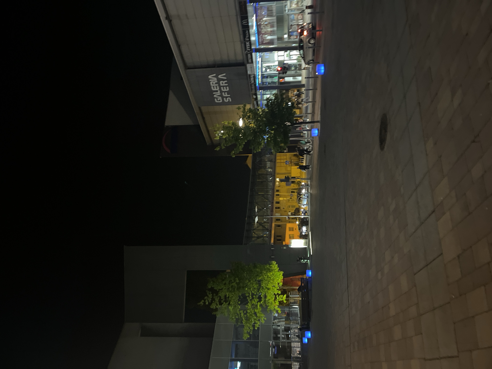 Here is a description of the image:
This is a night shot of an urban landscape. On the right is a brick-paved street illuminated by dim yellow light. On the left is a building that appears to be a shopping center named "Galeria Sfera." There are trees and illuminated blue cube lights along the street. Farther back, city lights and traffic are visible. The sky is dark.