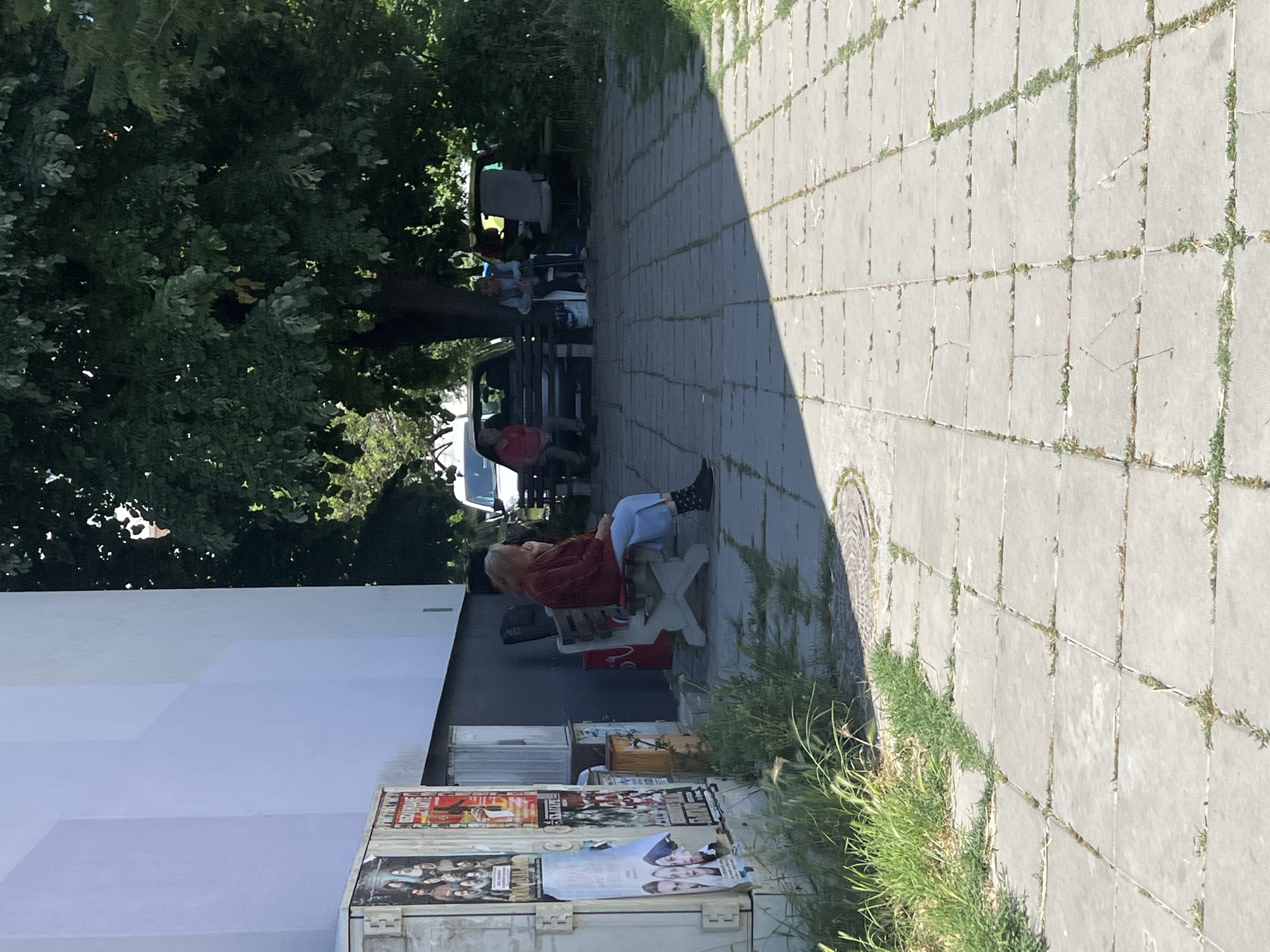 The image shows a scene on a sidewalk in Burgas, Bulgaria. The sidewalk is paved with large gray concrete blocks and has grass growing in the cracks. A woman is sitting in a white plastic chair on the sidewalk, wearing a red shirt, light blue pants, and black boots with white polka dots. Behind her, near the side of a light-colored building, are posters affixed to a structure, possibly a container or storage unit. Further down the sidewalk, under the shade of some trees, are a group of people sitting on benches and chairs. A car is parked nearby. The scene appears to be a public space where people are gathering and relaxing.