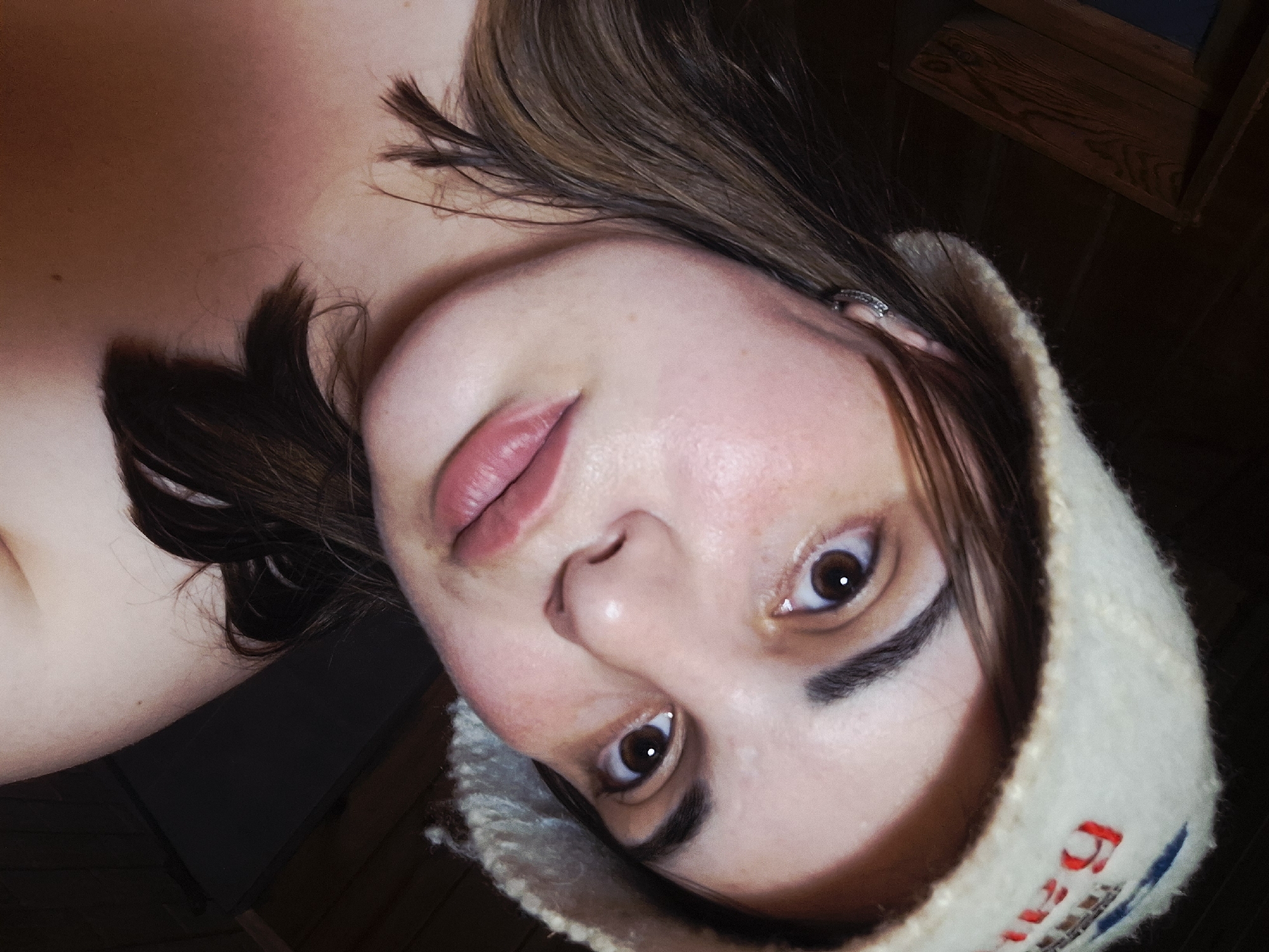 A close-up, angled, indoor shot shows a fair-skinned woman wearing a white knit hat with some red and blue lettering on the rim. Her gaze is directed upwards towards the viewer. She has brown hair with some strands falling across her face. The upper left side of the image shows a bare shoulder and the side of her neck. The background consists of dark, unidentifiable elements and a small section of wood paneling. The image was taken in Амзя, Russia.