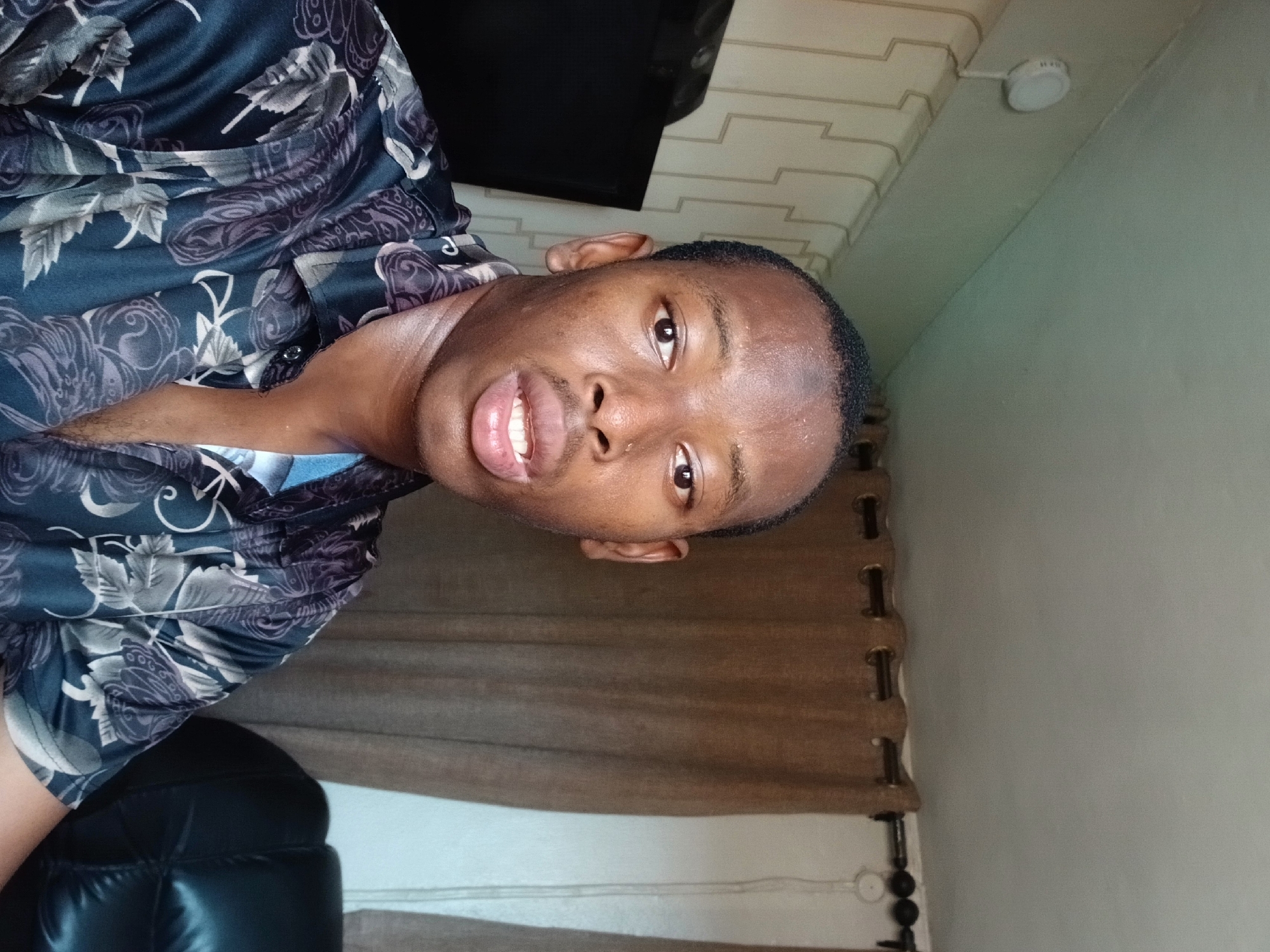 A man with short, dark hair, brown eyes, and dark skin is facing the camera with a slight smile. He is wearing a patterned, dark-colored button-down shirt. The background includes a wall with light-colored wallpaper, a dark-colored flat-screen television, and brown curtains. A ceiling light fixture is visible. The setting is likely indoors. There is no explicit indication this image was taken in Lagos, Nigeria.