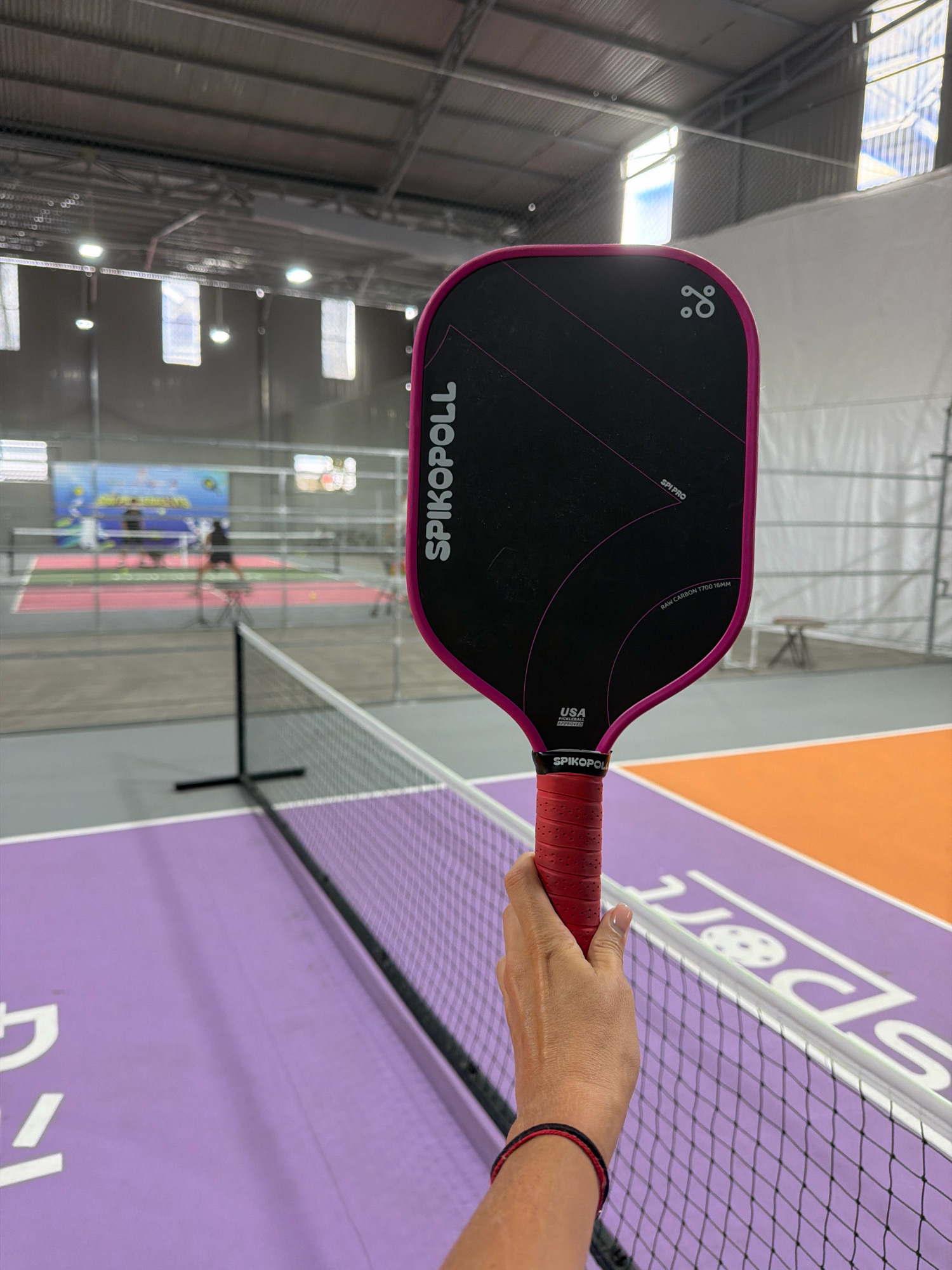 A person's hand, wearing a red and black bracelet, grips the red handle of a black pickleball paddle with a pink edge. The paddle is held in the foreground, obscuring part of an indoor pickleball court. The court has purple and orange sections, with a white net dividing them. In the background, other pickleball courts are visible, with players in silhouette. The facility appears to be an indoor sports complex with a metal roof and skylights. The overall scene depicts an indoor pickleball court setting. There is no indication of the city of Da Lat, Vietnam, in the image.