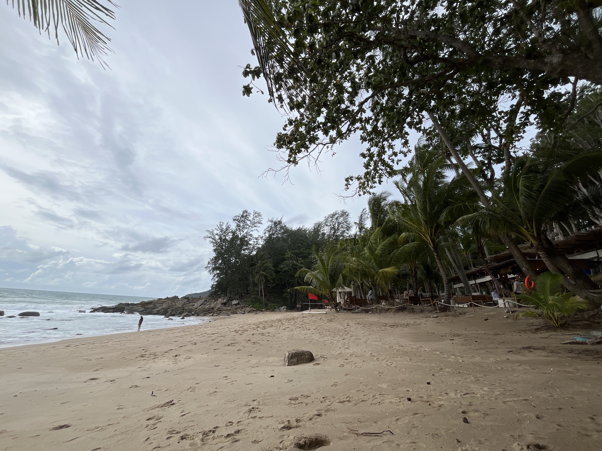 A wide, sandy beach in Choeng Thale, Thailand, faces a grey, overcast sky. Waves gently roll onto the shore, and scattered rocks are visible near the water's edge. A lone figure stands near the water, looking out towards the sea. Palm trees line the back of the beach, with some vegetation and a small resort or restaurant visible amongst them. The sand is marked with footprints and a few scattered stones and debris.