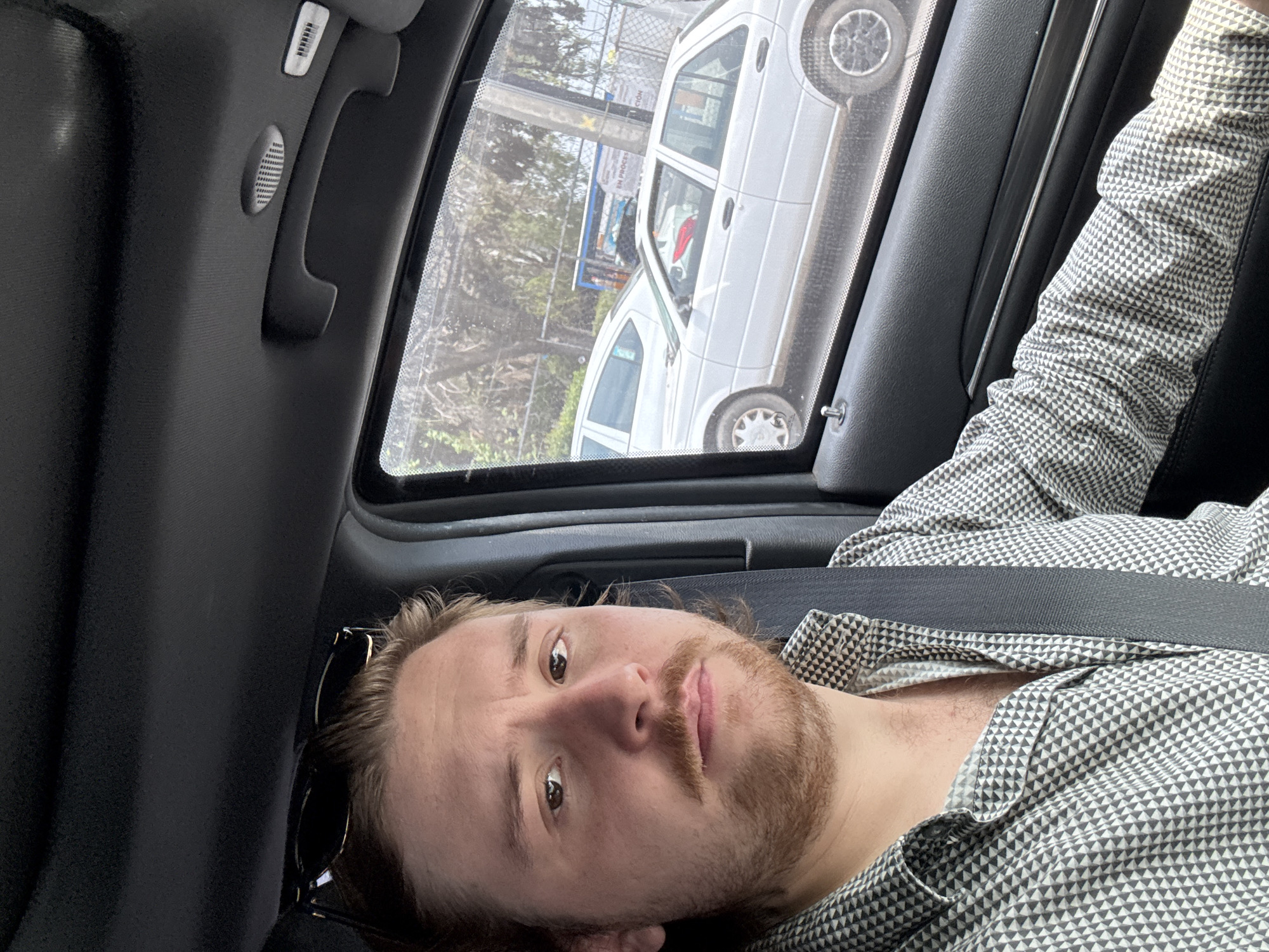 A man with brown hair and a light beard is lying in a car, wearing a patterned shirt and a seatbelt. Sunglasses are visible near his head. A white car is seen through the car window, and a sign reading "EN PROCESO" is partially visible. The location is likely Cuautitlán Izcalli, Mexico.