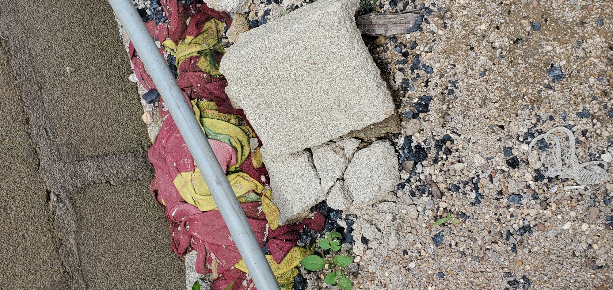 In an outdoor environment, possibly a construction site or a neglected urban area, a pile of red and yellow fabric is partially obscured by a grey metal pipe. The fabric rests on a ground surface composed of gravel, small stones, and dark debris, possibly charcoal or asphalt. In the foreground, a broken concrete block, light grey and textured, lies on the gravel. To the left of the concrete block, a few small green plants are growing. Further down in the gravel, a white mesh material is visible. The background consists of a textured, grey wall, possibly concrete, with a rough, uneven surface. The scene appears to be a still life of discarded materials and elements of nature in an urban setting.