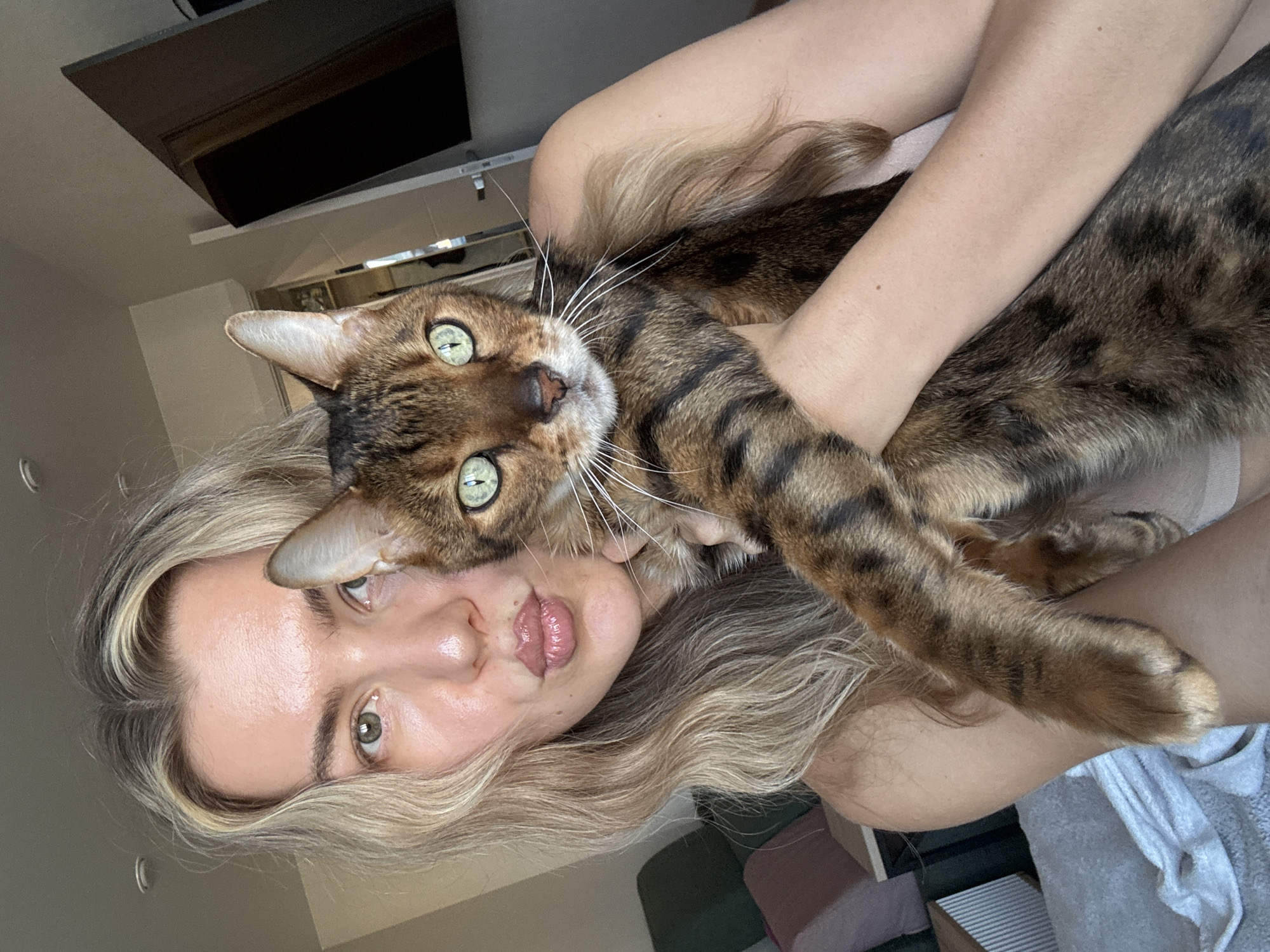 This image, rotated 90 degrees clockwise, depicts a woman holding a cat in what appears to be an indoor setting. The woman's head is positioned on the left side of the frame, with her body extending towards the right. She has long, wavy blonde hair that partially covers her shoulder and cascades around her face. Her complexion is fair, and her eyes, appearing green or hazel, gaze directly at the viewer. Her lips are slightly pursed in a common selfie expression. She wears a light-colored top, and a piece of grey fabric, possibly a blanket or sweatshirt, is visible near her right arm.
She is embracing a domestic cat, which looks like a Bengal or similar breed, recognizable by its distinctive brown and black spotted and striped fur pattern. The cat's face is prominent, positioned very close to the woman's head, and it also looks straight at the camera with striking, bright green eyes and long, white whiskers. Its paws are visible, resting on the woman's arm and shoulder as she holds it.
The background is softly focused and indistinct, suggesting an indoor environment, possibly a home. A dark, rectangular object, likely part of furniture or a shelf, can be seen in the upper left portion of the image. The lighting is soft and natural, indicating it is daytime. The scene conveys a moment of affectionate interaction between the woman and her pet. There is no visible text in the image.