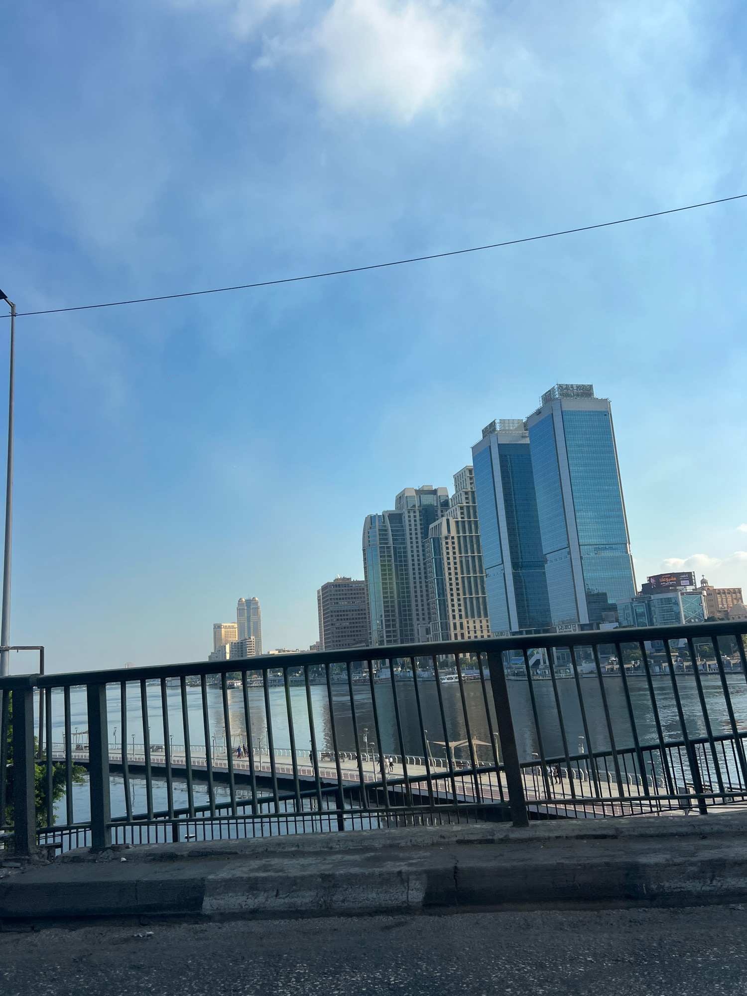 In a city scene, a body of water, possibly a river, reflects the sky and surrounding buildings. A dark metal railing in the foreground separates the viewer from a pedestrian walkway that extends over the water. On the walkway, a few individuals can be seen. Behind the walkway, modern glass-fronted skyscrapers rise against a clear blue sky with scattered clouds. The architecture suggests a contemporary urban environment. The perspective is from a slightly elevated position, looking across the water towards the city skyline.