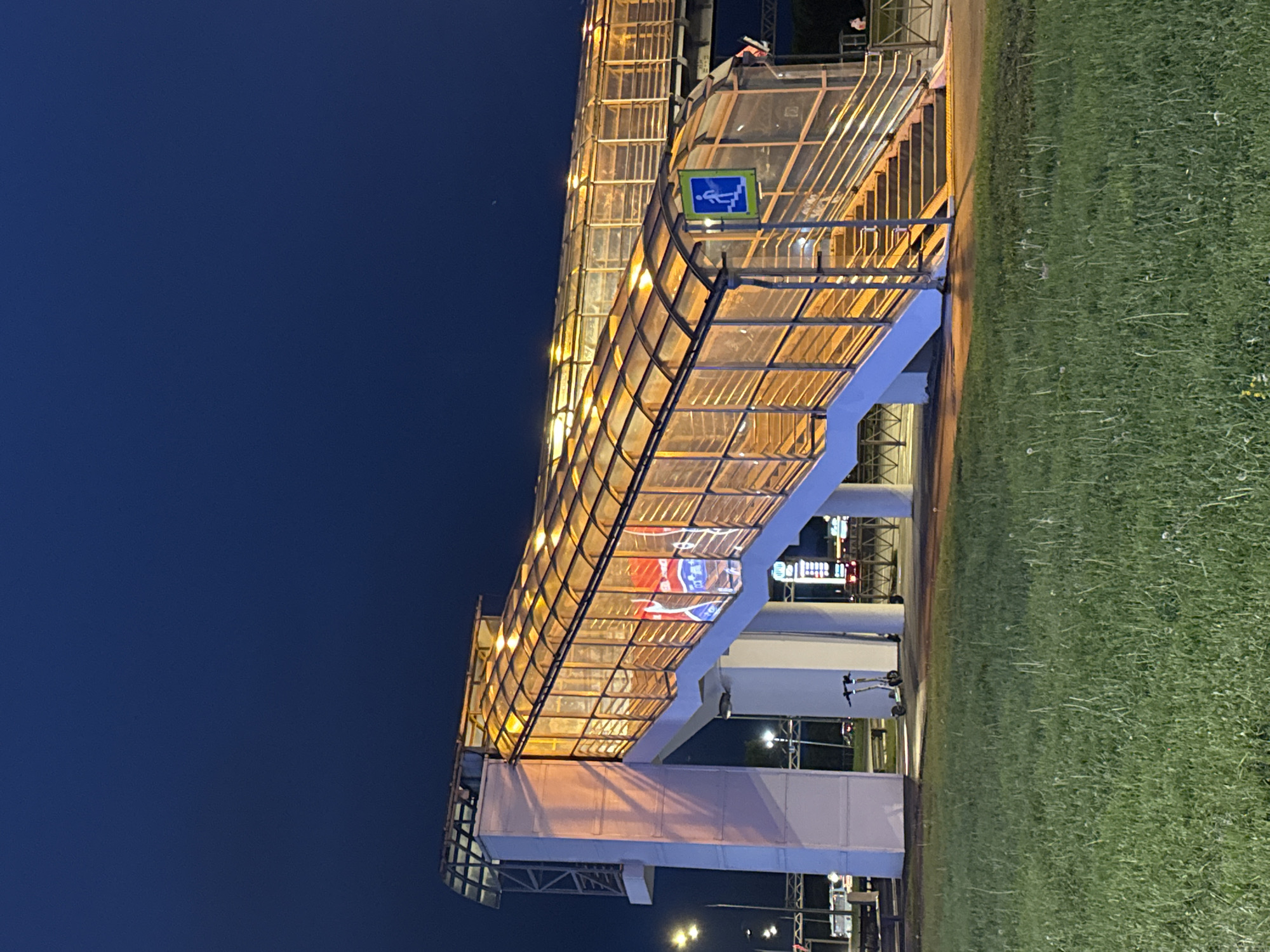 The image depicts a pedestrian overpass in Saint Petersburg, Russia, at night. The overpass is brightly lit, with a translucent roof and metal frame. A blue and white pedestrian sign is visible near the upper part of the overpass. Russian flags are draped inside the overpass. The sky is dark, and a green lawn borders one side of the overpass. Streetlights are seen in the background. A scooter is on the sidewalk by the overpass.