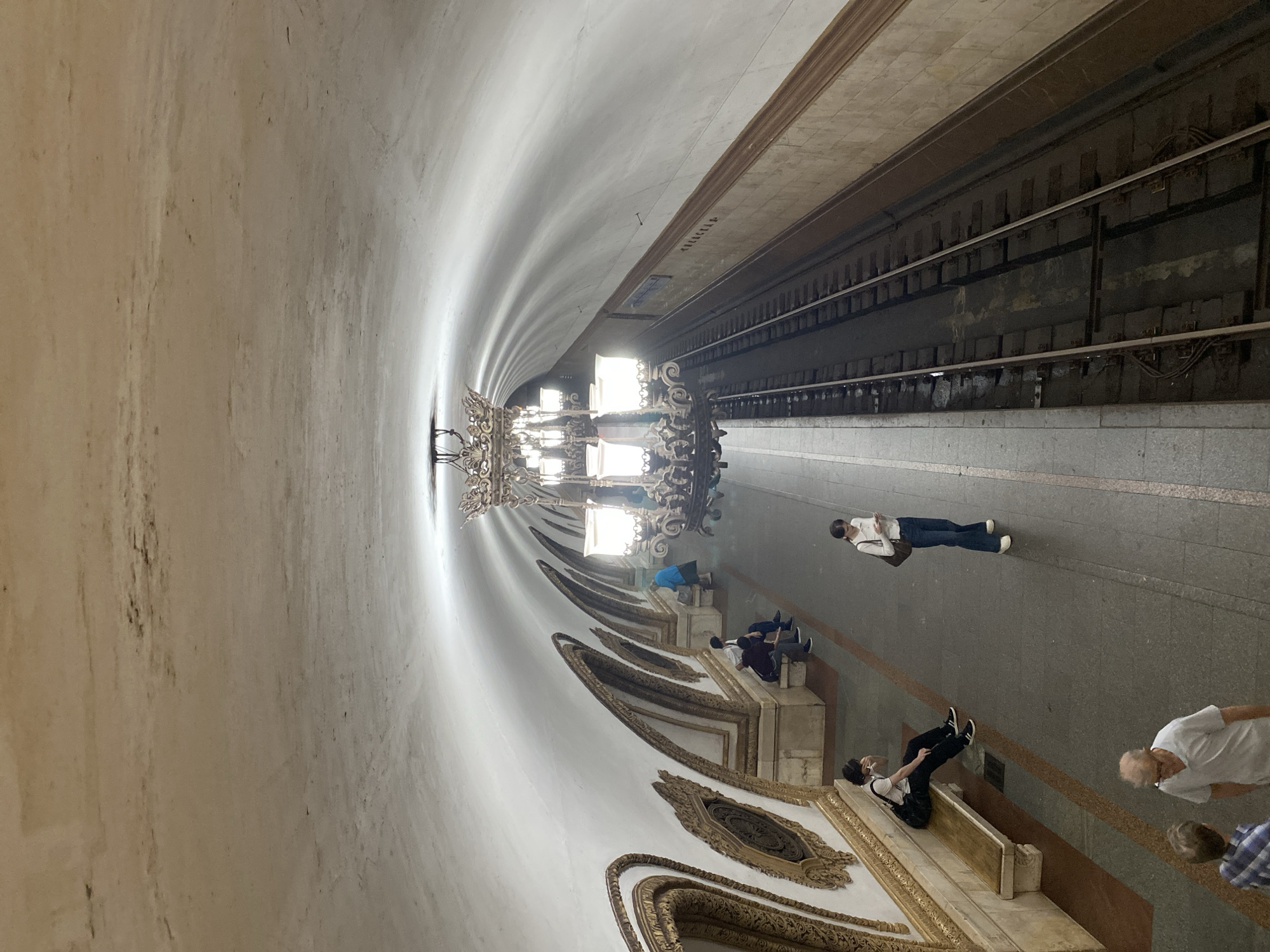 The image depicts a view from a high vantage point looking down onto a subway platform in Moscow, Russia. The station features ornate architecture with arched alcoves decorated in gold trim along the left wall. A large, ornate chandelier hangs from the vaulted ceiling, illuminating the space.
On the platform, several individuals are visible. A person is seated on a bench on the left, looking at their phone. Further down the platform, another individual stands, also looking at their phone. Two other individuals are standing together in the foreground, appearing to converse. In the distance, more people are seated on benches.
The subway tracks run along the right side of the image, disappearing into the tunnel. The platform is made of concrete, with yellow lines marking the edge. The walls of the station are a pale, textured material, with darker markings that suggest age or discoloration on the ceiling. The overall atmosphere appears to be one of quiet anticipation, typical of a subway station.