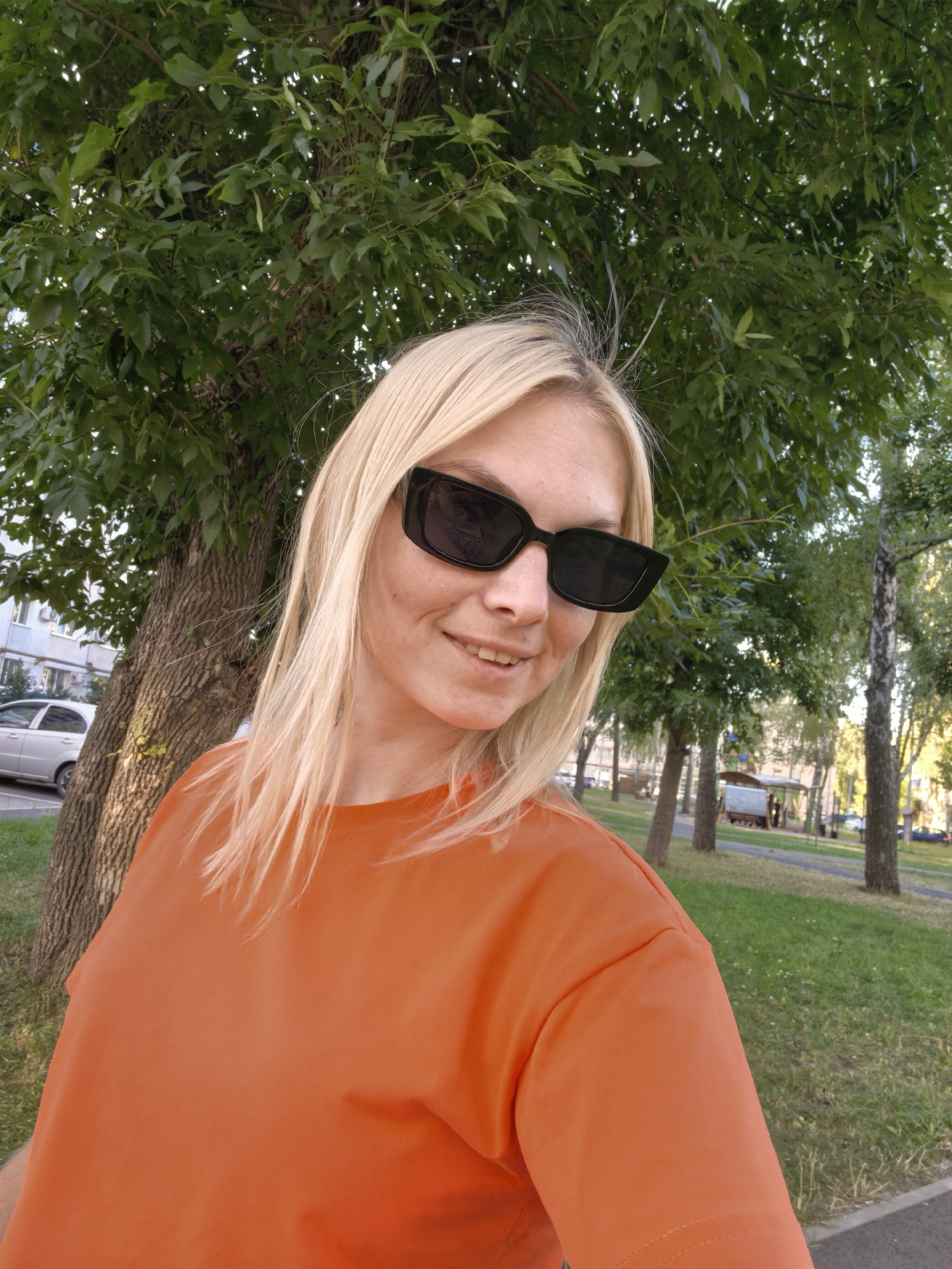 In Sterlitamak, Russia, a woman with blonde hair and sunglasses stands outdoors, facing the camera and smiling. She is wearing an orange t-shirt. Behind her, a tree with a rough bark trunk is visible, and further in the background, there is a grassy area with more trees and a pathway. A car is parked on the left side of the image. The scene appears to be a park or residential area on a clear day.