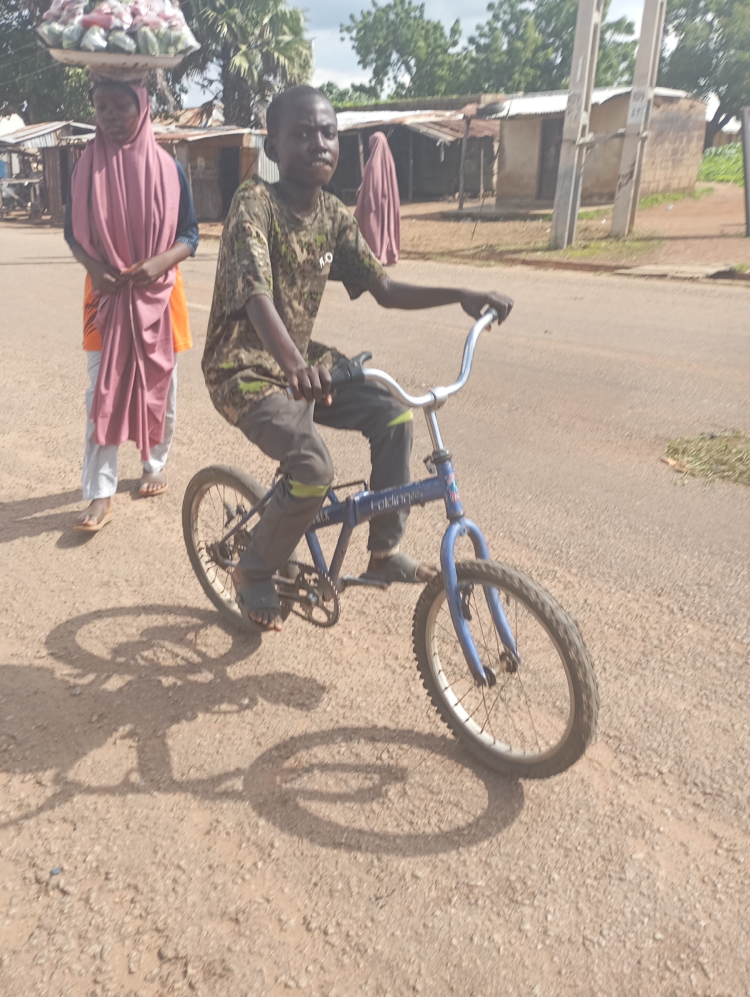 In Shendam, Nigeria, a young Black male, appearing to be of primary school age, rides a blue BMX-style bicycle on a dirt road. He wears a camouflage-patterned t-shirt and dark pants with a lime green accent stripe above the knee. His feet are clad in dark sandals. He is looking forward, with his hands on the handlebars. To his left and slightly behind him, a Black female, also appearing to be a child or young adolescent, walks. She wears a pink headscarf and a blue shirt, with an orange garment visible underneath. She is carrying a large tray laden with produce, possibly fruits and vegetables, on her head. The background features simple buildings with corrugated metal roofs, some appearing to be stalls or small shops, and a utility pole. Trees with green foliage are visible behind the structures. The ground is a dusty, reddish-brown dirt surface, and the shadows of the people and the bicycle are cast upon it, indicating a sunny day.