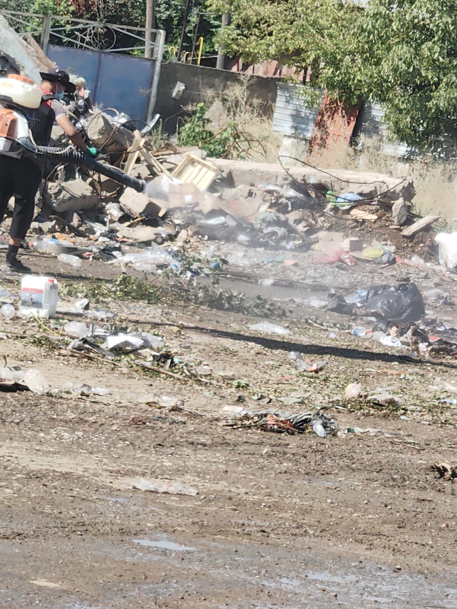 In Shymkent, Kazakhstan, a person wearing black pants, a black vest over a grey shirt, a black hat, a red bandana around their neck, and green gloves is operating a leaf blower. The leaf blower has a white tank and an orange engine. The individual is standing in a large pile of debris, including rocks, broken wood, plastic bottles, and black trash bags. The environment appears to be an outdoor area with a dirt ground, some vegetation, and a building in the background. The scene suggests a cleanup or demolition operation in a cluttered, possibly derelict, urban setting.