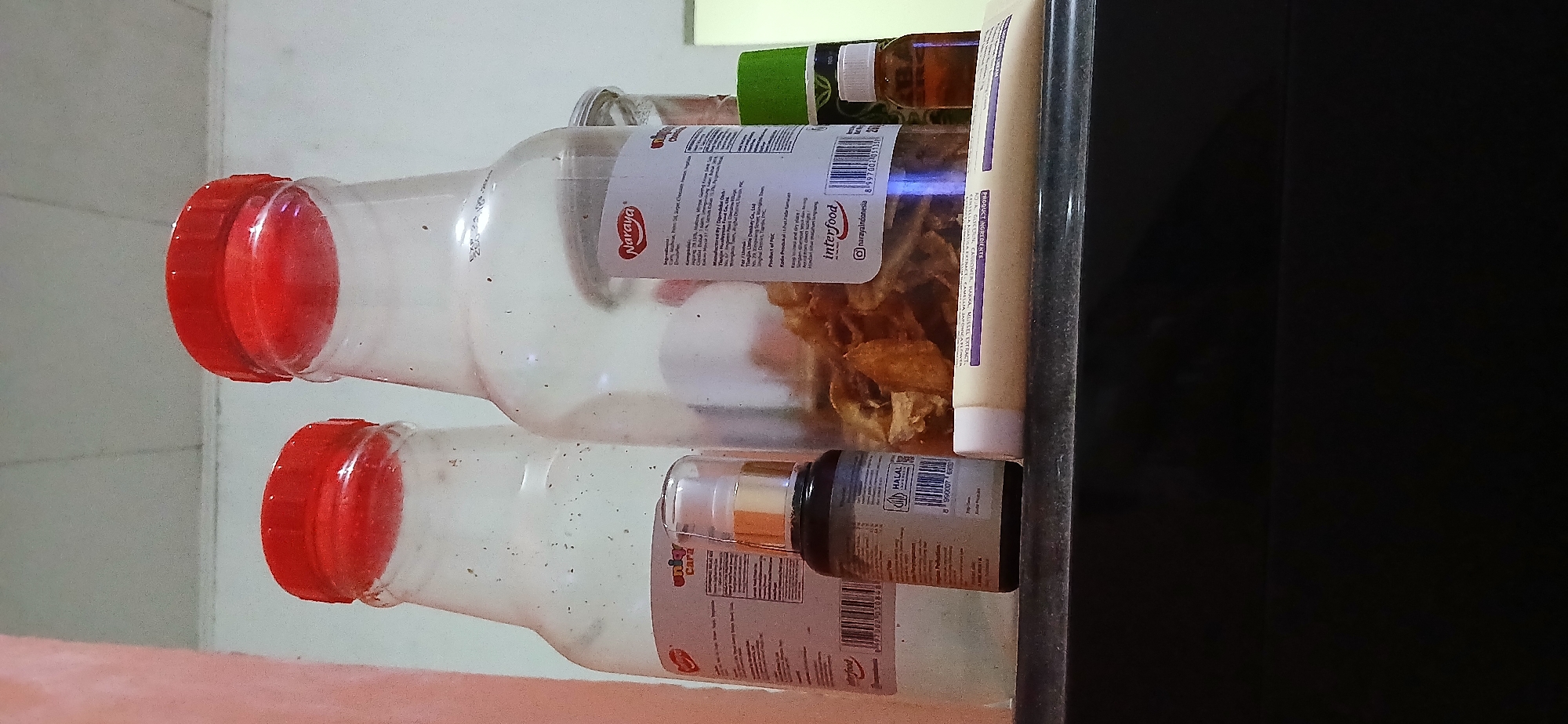 The image shows a shelf with several items, including two large plastic bottles with red caps, a small dark bottle with a gold pump dispenser, and a tube of cream. The larger bottles appear to be food containers, with labels visible. One label shows "Naraya" and ingredients listed in Indonesian. The smaller bottle has a label with a barcode and the word "HALAL" in a green circle. The tube of cream has text that includes "CERATIN" and "EXTRACT". The shelf appears to be made of a dark material, possibly wood or laminate. The background is a light-colored wall. The overall impression is of a cluttered shelf in a home or small shop. There is no indication of the location being in Samarinda, Indonesia.