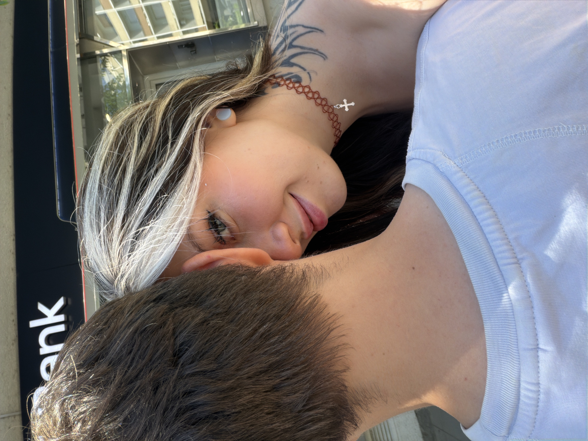 A close-up shot shows a woman resting her face on a man's head. The woman has blonde and brown hair, fair skin, a tattoo on her neck, a brown choker with a silver cross, and what appears to be a wireless earbud in her ear. The man has dark brown hair. He is wearing a white T-shirt. The image is taken outdoors. A bank building can be seen in the background. Without additional contextual information, it is difficult to confirm the precise location.