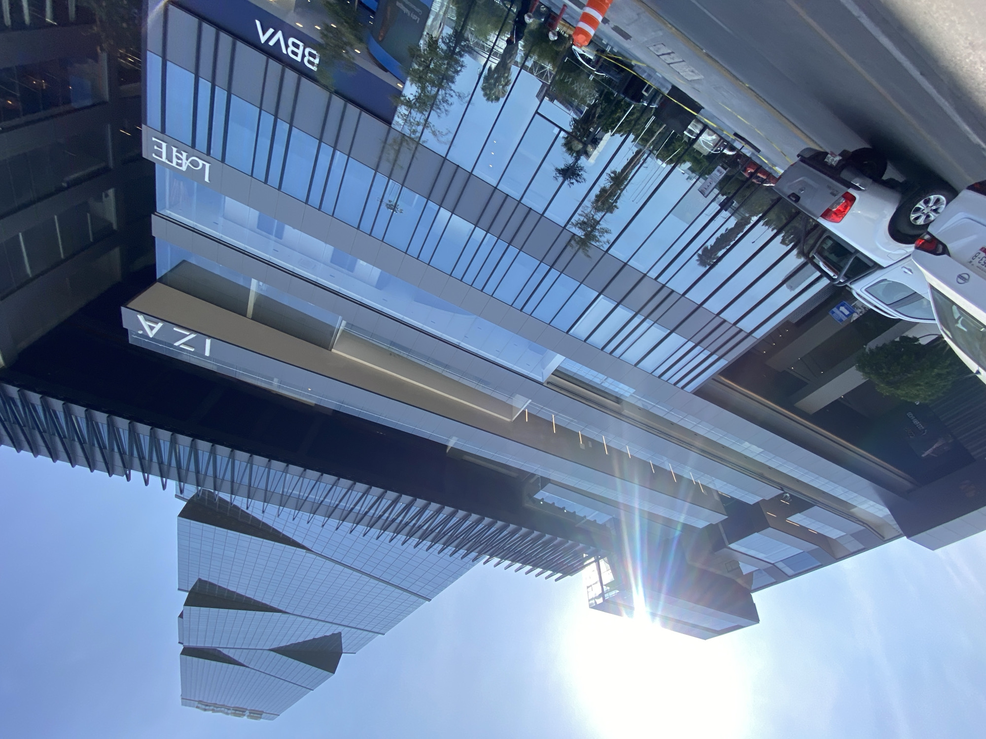The image shows a modern office building complex on a clear, sunny day. The building features multiple glass panels that reflect the sky and surrounding trees. The names "IZA", "LOFTE", and "BBVA" are displayed on different parts of the building. A street with parked cars and construction cones is visible at the top. A taller skyscraper-like building stands out against the blue sky, creating a layered architectural landscape. The location is in the city of Unknown, Unknown.