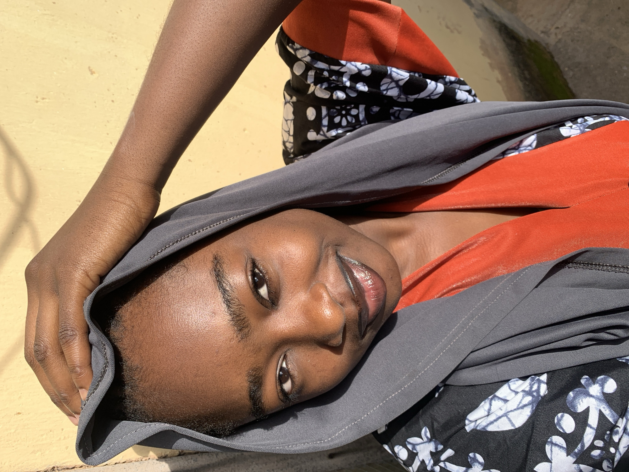 In an outdoor, sunlit environment, a young Black woman is captured in a close-up portrait. She is wearing a gray hijab and a vibrant orange top layered with a black and white patterned garment. Her right hand is raised, touching the top of her head, which is covered by the hijab. The background consists of a textured yellow wall, suggesting an urban or residential setting within Rafin Par, Nigeria. The lighting creates strong shadows and highlights, emphasizing the contours of her face and the textures of her clothing and the wall.