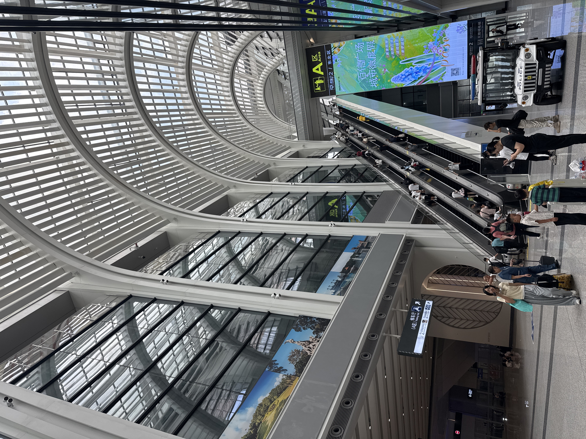 The image depicts the interior of a modern transportation hub, possibly a train station or airport, in Baiyun District, China. The architecture is characterized by a soaring, arched ceiling with a slatted roof, allowing natural light to filter through. Large glass windows line the walls, reflecting the interior and the sky above.
A prominent escalator, filled with people traveling in both directions, ascends through the center of the space. The escalator is flanked by large digital screens displaying vibrant advertisements. To the right of the escalator, a small, white electric vehicle with "POLICE" labeling is parked, suggesting a security presence.
In the foreground, several individuals are visible. A woman in a light-colored t-shirt and grey pants is walking with a suitcase. Other people are standing and talking, some with luggage. The floor is polished and reflective, contributing to the sense of spaciousness.
Signs are visible in both Chinese and English, indicating directions and information for passengers. One sign points to "Area A" and "Waiting Hall". The overall impression is one of a busy, contemporary public space designed for efficient transit.