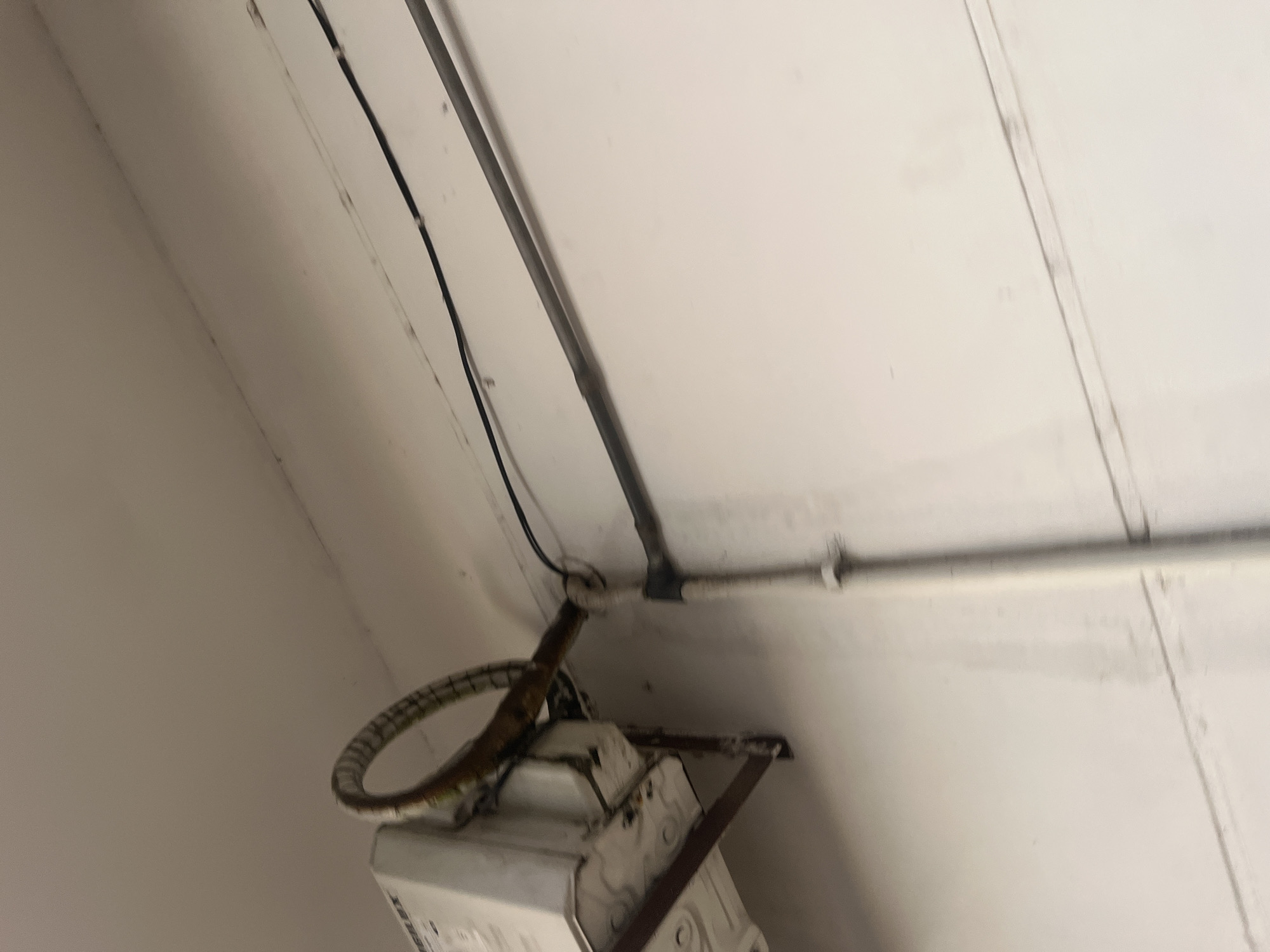 The image shows a close-up of an outdoor wall, likely made of concrete, painted white. A white electrical box is mounted on the wall by a metal bracket. A flexible, ribbed pipe with a dark interior curves from the electrical box towards the right side of the image. Below this, a dark electrical conduit runs horizontally across the wall, secured by metal straps. A vertical grey pipe, also secured by straps, runs down the wall to the right. The scene appears to be in an industrial or utility area, possibly a back alley or service area. The lighting suggests daytime. No people are visible. The location is described as being in Santa Ana, Venezuela.