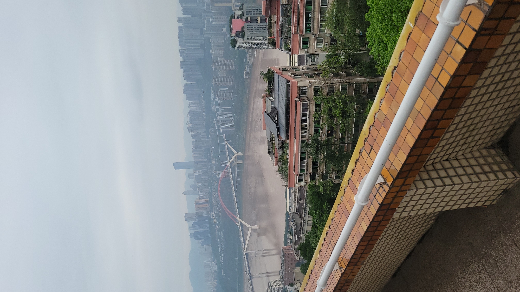 The image presents an urban scene of a city from an elevated perspective. A river flows through the cityscape with a bridge in the foreground, suggesting a potential infrastructure element. The architectural landscape features a dense arrangement of buildings, giving a sense of a populated area. The weather appears overcast, potentially affecting visibility. A section of the building's structure is visible in the foreground, showing construction or architectural detail with brown and white tiles and a white pipe.