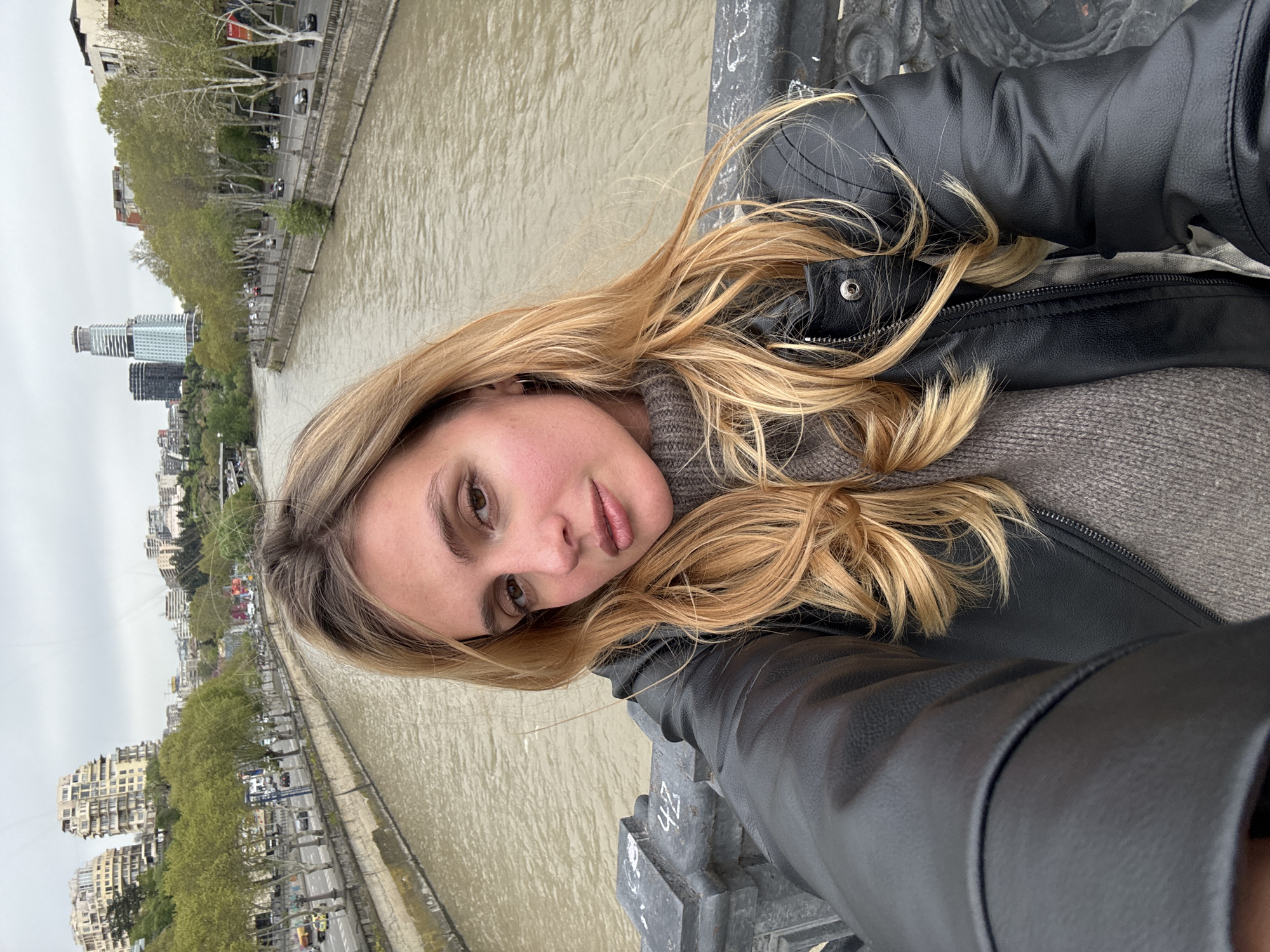 A young woman with long, blonde hair takes a selfie on a bridge in Tbilisi, Georgia. She wears a black leather jacket over a gray sweater. Behind her is a wide, muddy river lined with trees and buildings. In the distance, modern high-rise buildings dot the Tbilisi skyline. The sky appears overcast. The woman leans slightly over the bridge's railing, taking a picture. The bridge has metal railings.