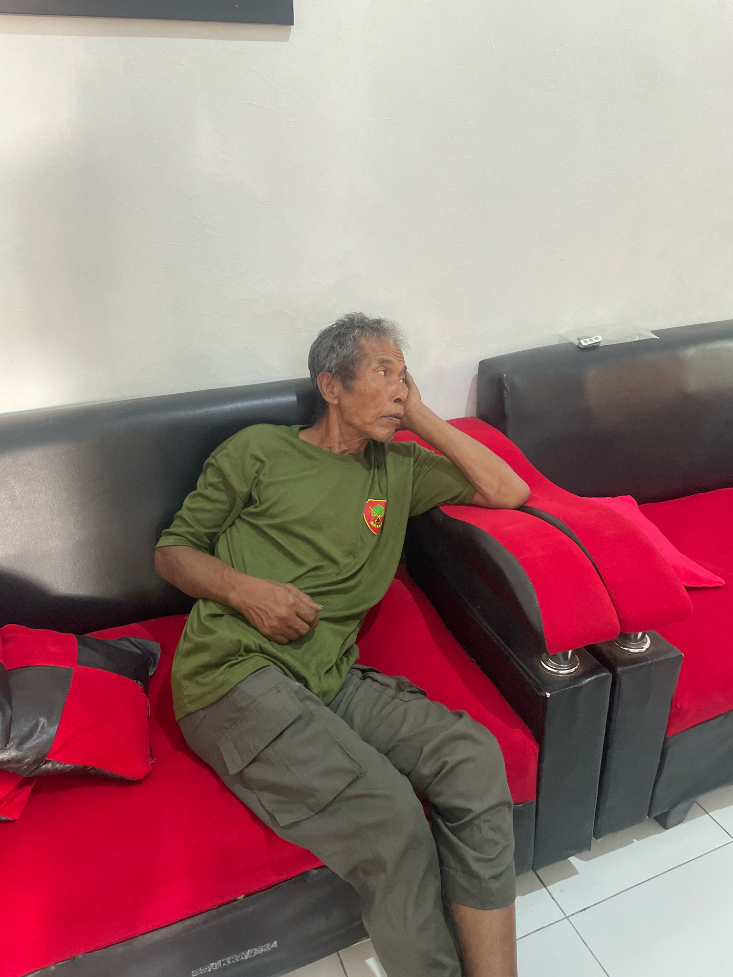 An elderly man with gray hair and wrinkled skin is sitting on a red couch. He is wearing a green t-shirt and gray cargo pants. The couch is a sectional, with black arms and red cushions. There is a pillow on the left side of the couch that is red and black. The man is leaning his head on his hand, looking off to the side. The background is a white wall. The image appears to have been taken indoors, likely in a home. The location in Kagungan Ratu, Indonesia, is not visually apparent from the image.