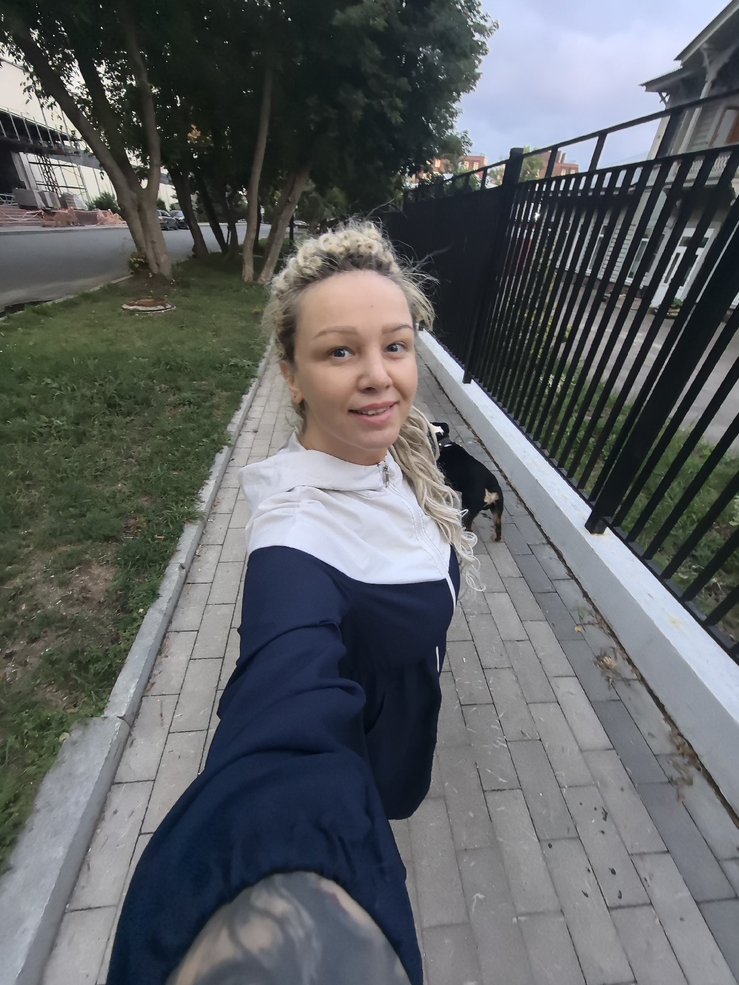In this full-body selfie taken from a low angle, a woman with blonde dreadlocks is walking on a paved sidewalk. She is wearing a dark blue and white hooded jacket. A small black dog with brown markings on its legs is walking behind her, to her right. The sidewalk is bordered by a strip of grass on the left and a black metal fence on the right. Beyond the fence, a building with windows and a railing is visible, along with some trees. The sky is overcast, suggesting it might be evening or a cloudy day. The scene appears to be in an urban or suburban environment. The woman's left arm is extended, holding the camera.
