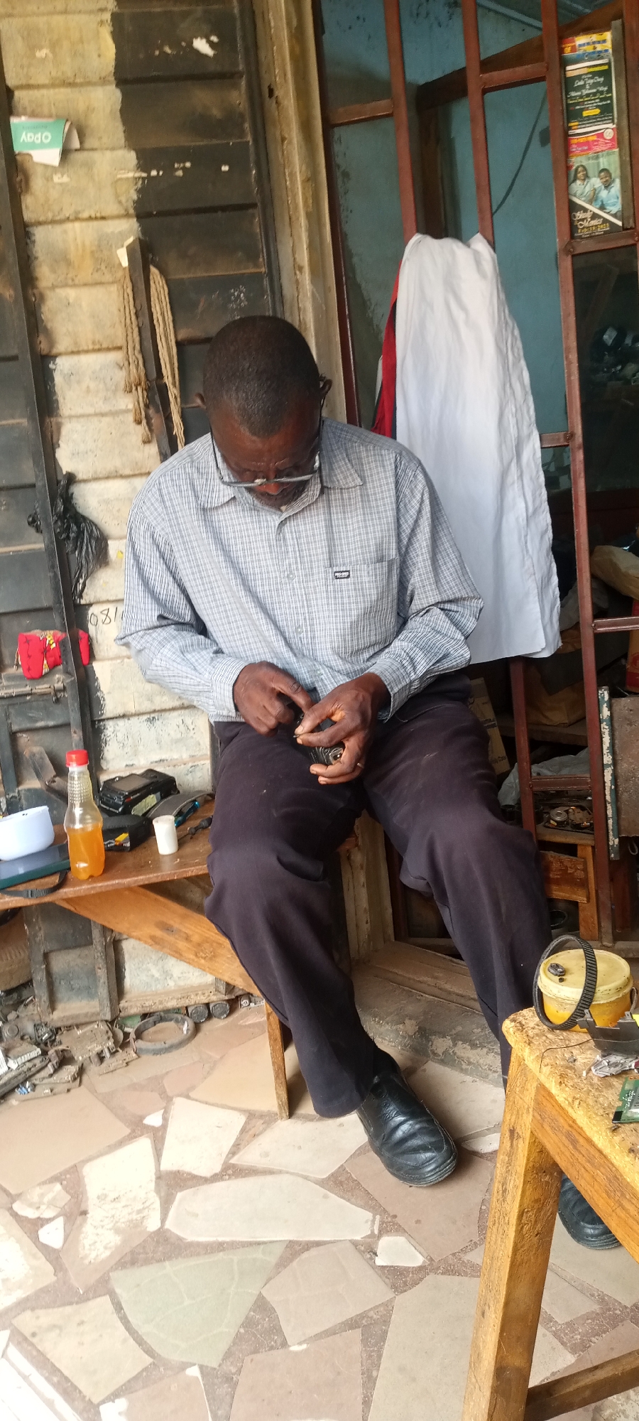 In Jos, Nigeria, an elderly man with glasses is seated on a wooden bench, meticulously working on a small, dark object with his hands. He wears a light gray plaid shirt and dark pants, with black leather shoes on his feet. The setting appears to be a workshop or small business, judging by the various tools and components scattered on the bench beside him and on the floor. A bottle of liquid is visible on the bench next to various tools, with a white towel hanging behind him. Two posters are visible on the frame behind him. The floor is tiled with light-colored geometric patterns.