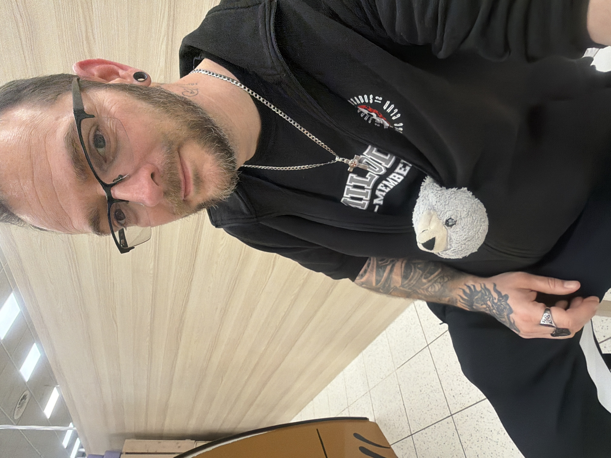 A man with glasses, a beard, and tattoos is indoors with a table and tiled flooring visible. The man is wearing a black vest, a black shirt with white lettering, and a silver chain with a cross. He has a small stuffed toy attached to his shirt. His left hand, with a ring and tattoos, rests on his right hand. The photo was taken in Saarburg, Germany.