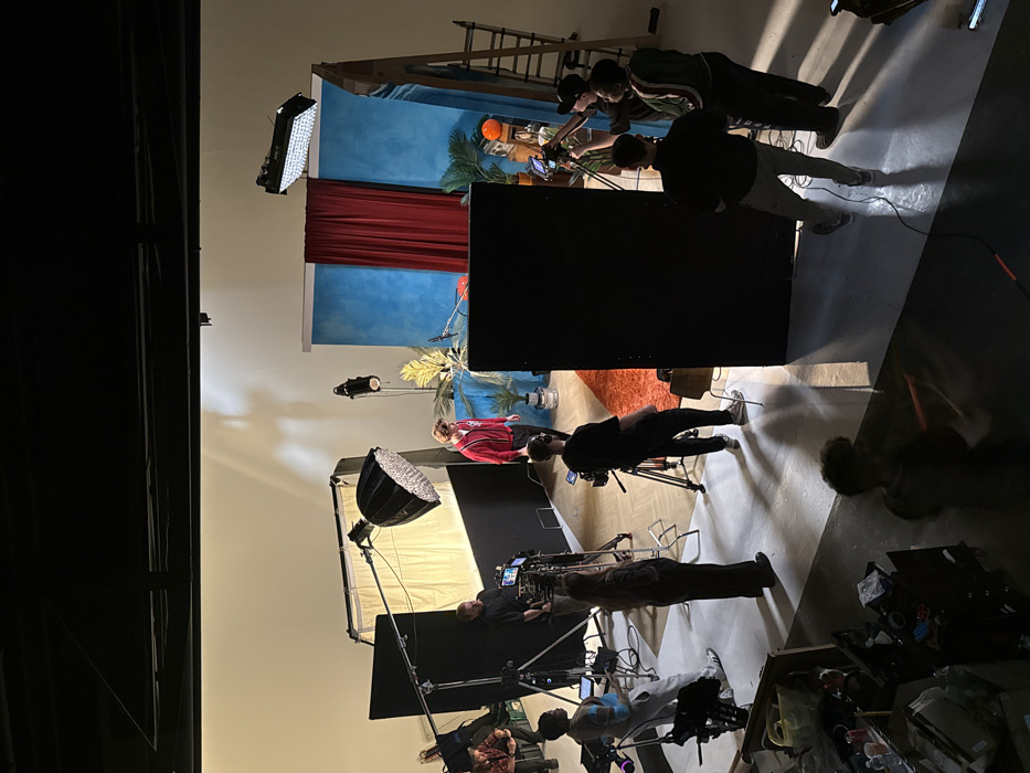 An indoor film or photoshoot production is depicted within a large studio space. The specific location in the city of London, United Kingdom, cannot be confirmed from the visual information provided.
The central focus is a male subject, wearing a red and white striped long-sleeved top and light trousers, standing on an orange rectangular rug. He faces towards the right, positioned in front of a backdrop featuring a textured blue section with a red curtain partially drawn across it. Various potted plants, including palm-like foliage, are arranged on either side of the subject. An orange radio and a small plant pot are visible on the right side of the set.
Approximately ten individuals are present, actively engaged as crew members. Key personnel include:
* A male operating a large video camera on a tripod to the left of the subject.
* A female standing near this camera, observing the set.
* A male positioned behind a large black panel on the left, holding a small monitor.
* A group of three males on the far right, one operating a camera on a tripod, another viewing a screen, and the third observing.
* A male seated on a stool in the far left background.
* A male in the foreground, blurred due to motion.
Lighting equipment includes a large parabolic softbox on a stand to the left, a rectangular LED panel light suspended above the backdrop, and a spotlight on a stand. Multiple black panels are strategically placed for light control. Three professional video cameras on tripods are visible. A wooden ladder leans against the backdrop structure on the right.
The studio environment features light-colored walls, a dark ceiling with visible infrastructure, and a floor comprised of light wood parquet in some areas and solid light grey in others. Artificial lighting casts distinct shadows across the floor and walls. The foreground area contains various production equipment and boxes, indicative of an active workspace.