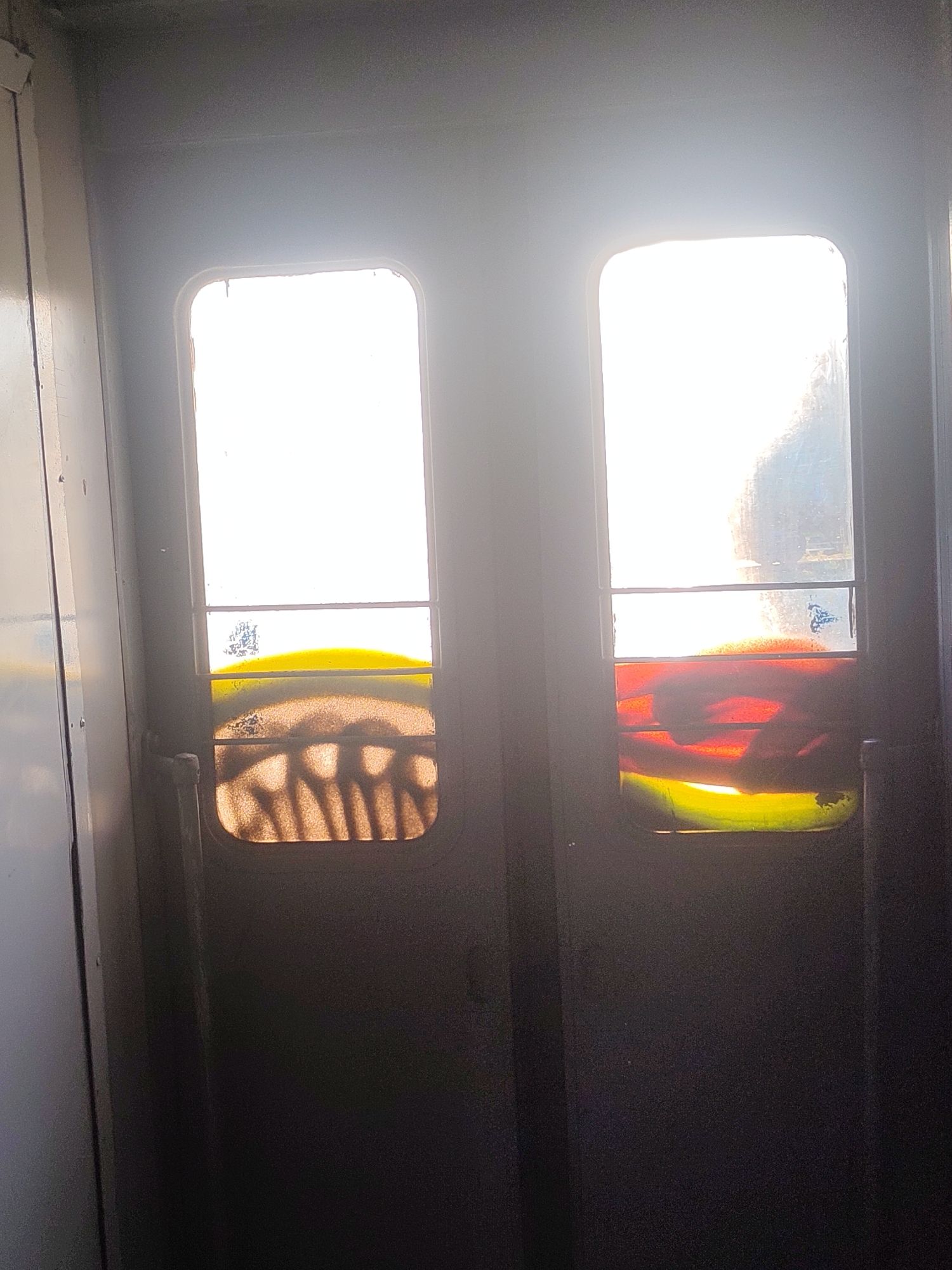 The image depicts a close-up view of two glass panels within a dark, metallic frame, likely a door or partition. The panels are illuminated from behind by a bright, white light, creating a strong silhouette effect. Each panel is divided horizontally by a thin metal bar. The lower portion of the left panel features a swirling pattern of brown and amber hues, resembling dust or sand trapped between the glass. The lower portion of the right panel displays vibrant, abstract streaks of yellow, red, and green colors, suggesting colored glass or some other material applied to it. The overall environment appears to be indoors, possibly within a public transportation vehicle or a similar structure, given the utilitarian nature of the framing and the presence of distinct panes. No specific landmarks or details are visible that would place this scene in Kyiv, Ukraine.