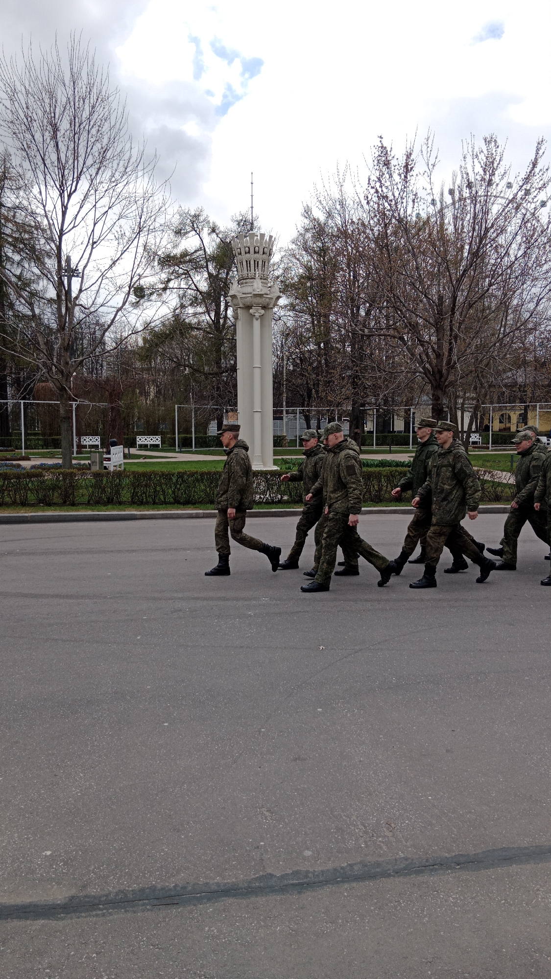 A group of uniformed soldiers march across a street in Moscow, Russia. The soldiers are wearing camouflage patterned uniforms and black boots. They are walking in a line, with the first soldier slightly ahead of the others. In the background, there is a park with trees, a white decorative column, and a building with a fence. The sky is overcast. The street is paved and appears to be lightly traveled.