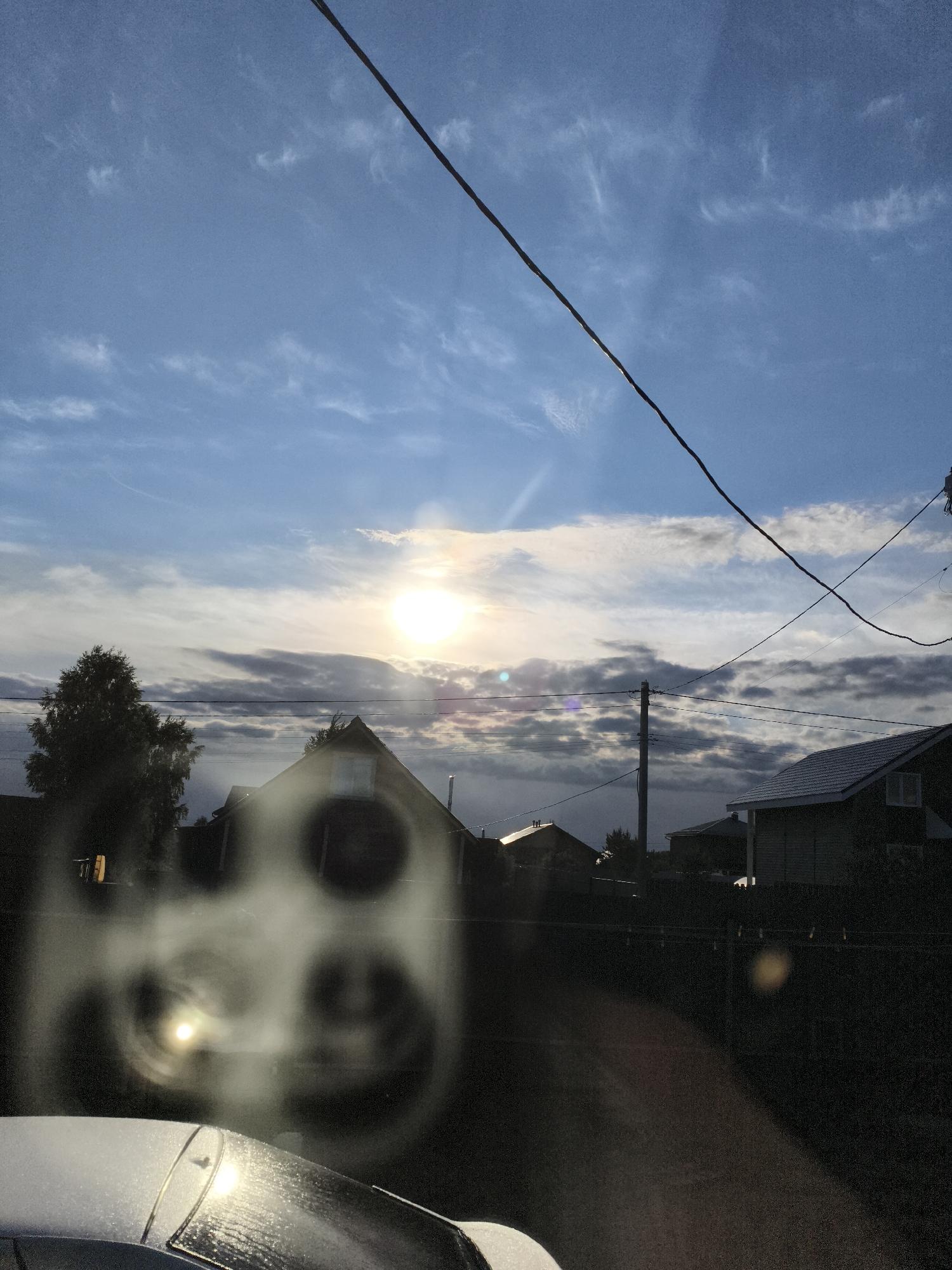 The image presents a tranquil, rural or suburban scene under a partly cloudy sky, likely captured during sunrise or sunset. A bright sun, positioned slightly right of center, dominates the upper middle section, casting lens flares and distinct circular reflections across the foreground, suggesting the photo was taken through a window or glass surface.
The sky transitions from a lighter blue with wispy clouds at the top to denser, darker clouds along the horizon. Several dark power lines traverse the sky, with one prominent line cutting diagonally across the upper right. A utility pole stands upright near the center-right, further indicating a developed area.
Along the horizon, dark silhouettes of houses and trees are visible against the brightening or fading light. Notable architectural features include an A-frame roof on a house to the left and a white-framed window on a building to the far right. These structures suggest a quiet, residential setting.
The bottom left of the foreground reveals the top portion of a car, including its hood and windshield, which appear covered in frost or dew, reflecting the sun's light. This detail, combined with the quality of light, points to either an early, possibly cold, morning or late evening.
No activity or people are visible in the static scene, and there is no discernible text. The overall impression is one of peaceful observation of the natural light and surroundings in Русский Вожой, Russia.