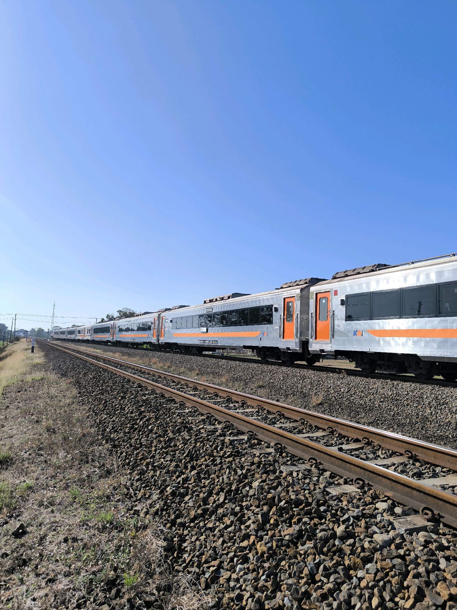 An exterior, eye-level, medium shot captures a long, silver and orange passenger train moving along a track on a clear day in Karanganyar, Indonesia. The train is to the right of the frame and moves from left to right. The track is in the foreground and is surrounded by gravel. The train is composed of several cars, each with orange doors and rectangular, dark windows. The sky is a clear, bright blue. In the distance, there are some trees and utility poles. The overall scene is one of travel and open space.