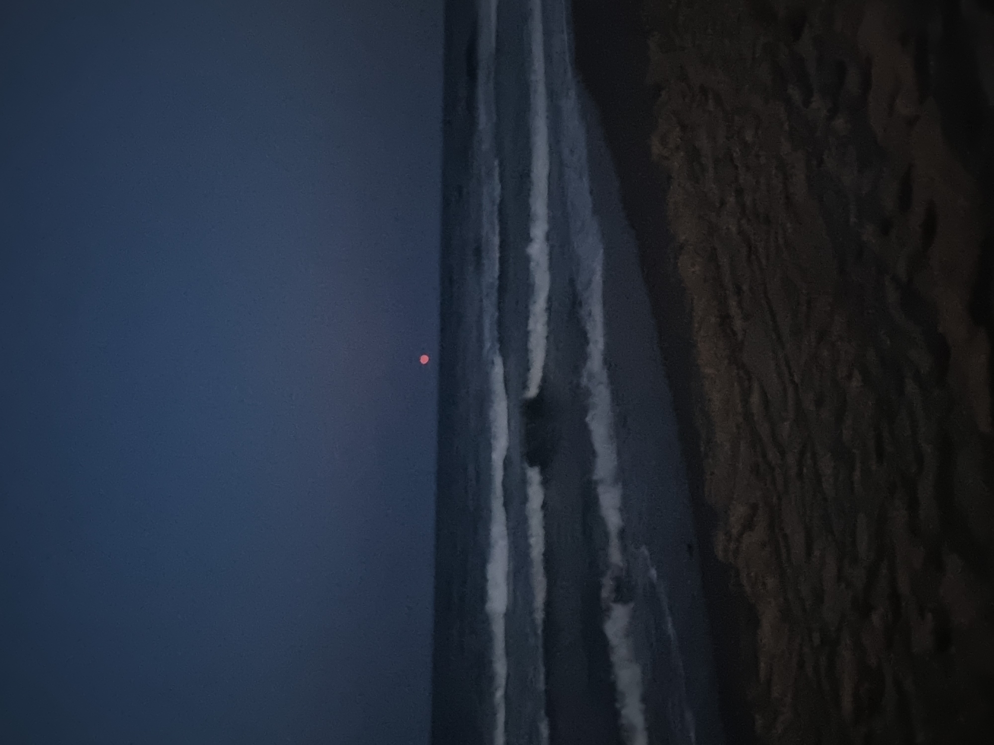 The image shows a nighttime beach scene in Tavernes de la Valldigna, Spain. The sky is a deep, dark blue. A small, reddish-pink moon is visible in the sky above the horizon. The sea is dark, with white-capped waves rolling towards the shore. The beach in the foreground is dark and appears to be made of sand, with some texture suggesting ripples or footprints. There are no people or animals visible in the image.