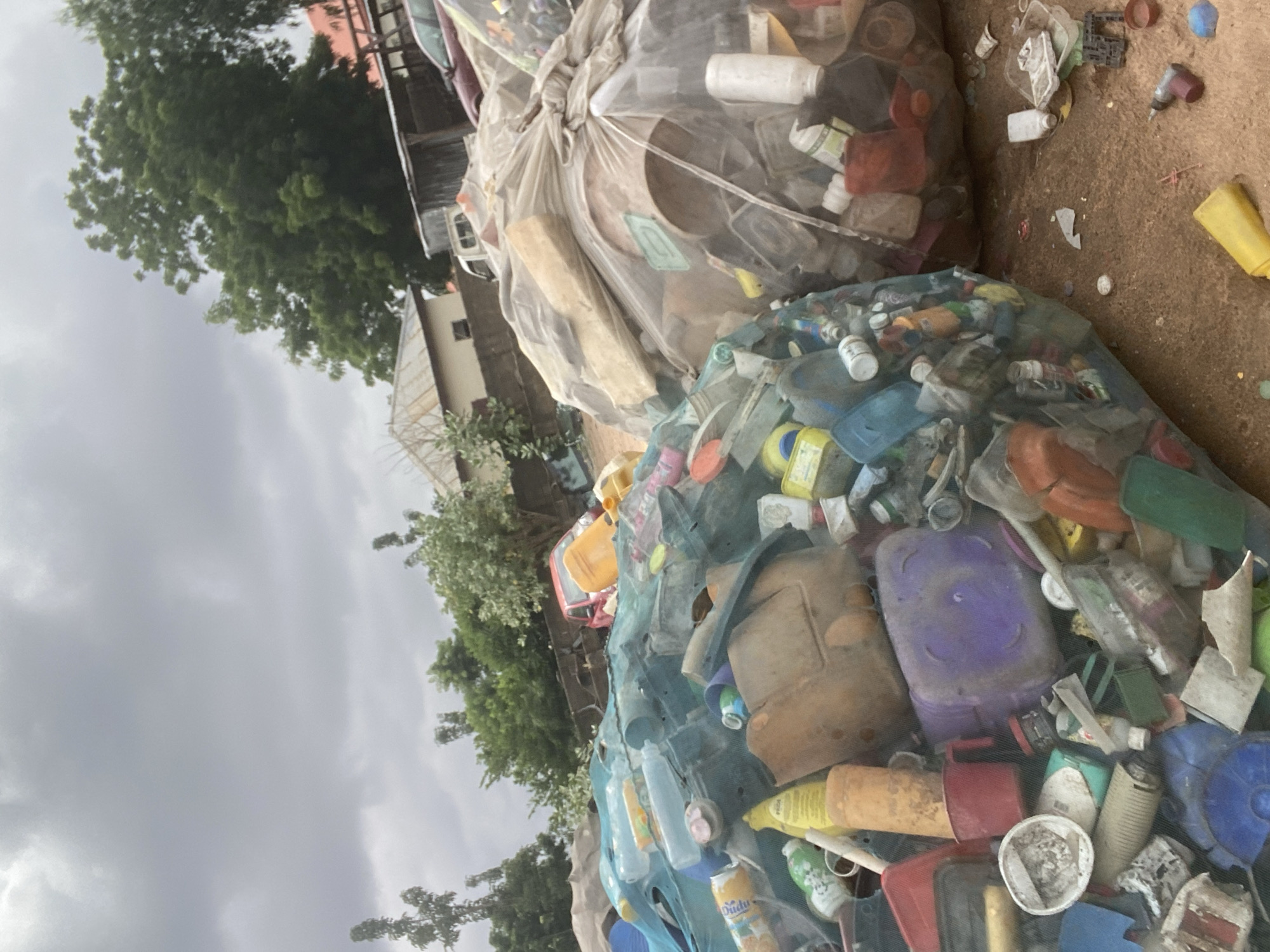The image depicts a scene of accumulated plastic waste at an open-air recycling or disposal site in Gbaza, Nigeria. Two large, translucent bags are filled with various plastic containers, bottles, and miscellaneous plastic items. The bag on the left is partially covered by a green mesh netting, revealing a mix of colorful plastic waste, including bottles, jugs, and containers. The bag on the right is predominantly filled with clear and white plastic items, including bottles and what appear to be larger plastic containers.
The ground beneath the waste bags is a dusty, reddish-brown earth, littered with scattered pieces of plastic debris, including a yellow plastic tube, a drill, and bottle caps. In the background, trees with dense green foliage are visible, along with some low-rise buildings with corrugated metal roofs. A small red car is also visible partially submerged in the waste, and a white utility vehicle is parked behind the structures. The sky is overcast and grey, suggesting a cloudy day. The overall impression is one of a neglected or overwhelmed waste management area.