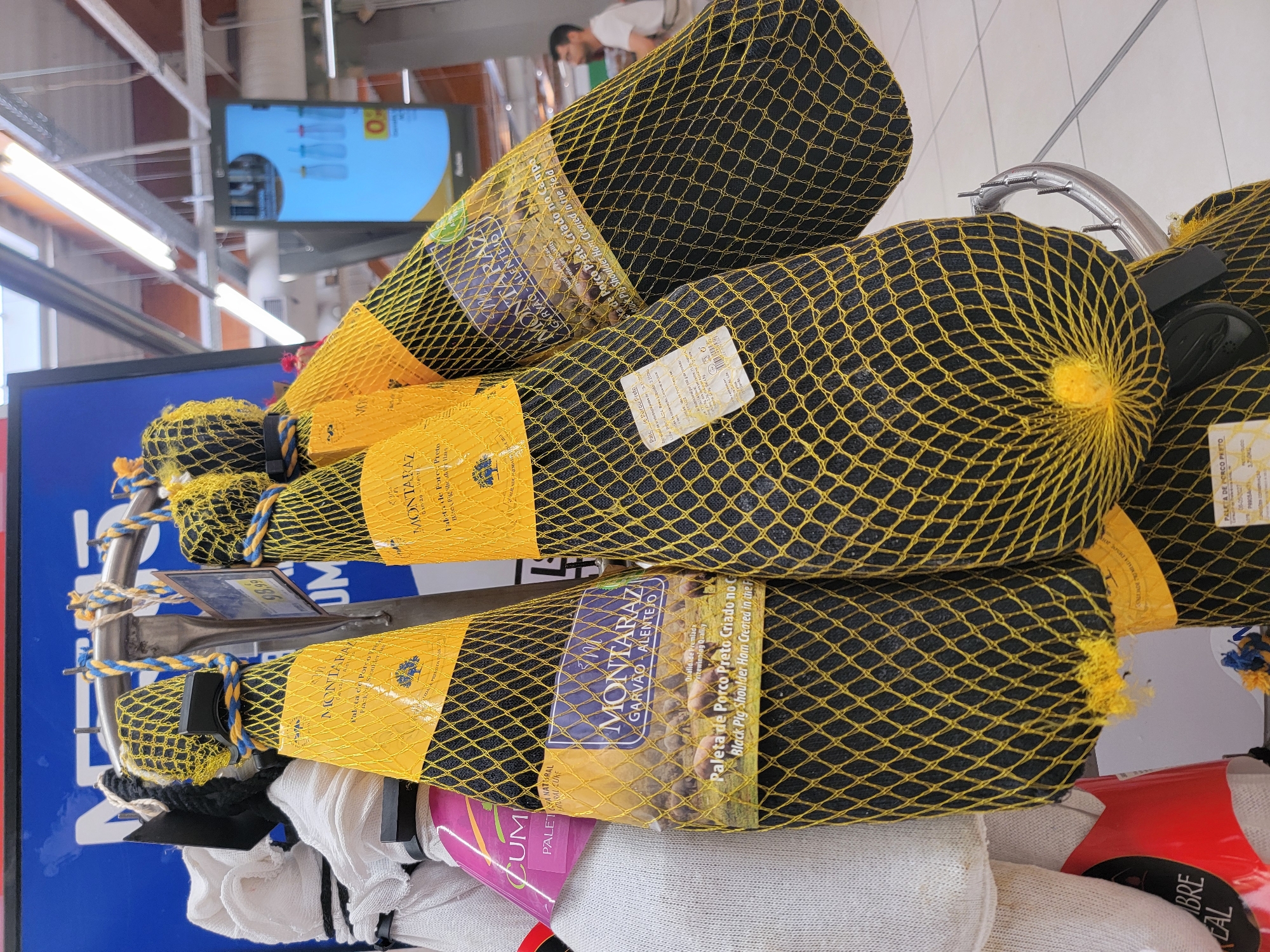 In a retail environment, possibly a supermarket in Faro, Portugal, several whole hams are displayed. The hams are dark in color and encased in black mesh netting with a yellow criss-cross pattern. Each ham has a yellow label attached, featuring the brand name "MONTARAZ" in prominent lettering, along with the location "GARVÃO - ALENTEJO." Below the brand name, there's a depiction of pigs grazing in a field and text in Portuguese and English: "Paleta de Porco Preto Criado no Campo / Black Pig Shoulder Ham Created in the Field." One label also specifies "Qualidade Premiada / Rewarding Quality." The hams are hung from a metal rack, with blue and yellow ropes tied around the tops of some of the hams, securing them. In the background, a digital screen displays images of baby bottles and text that appears to be an advertisement or information display. The lighting suggests an indoor setting with fluorescent fixtures visible on the ceiling. One person, a man with short hair and glasses wearing a white t-shirt, is partially visible in the background, looking towards the right side of the frame. Another white object, possibly another product or a display, is visible on the far left, with a pink label that reads "CUM..."