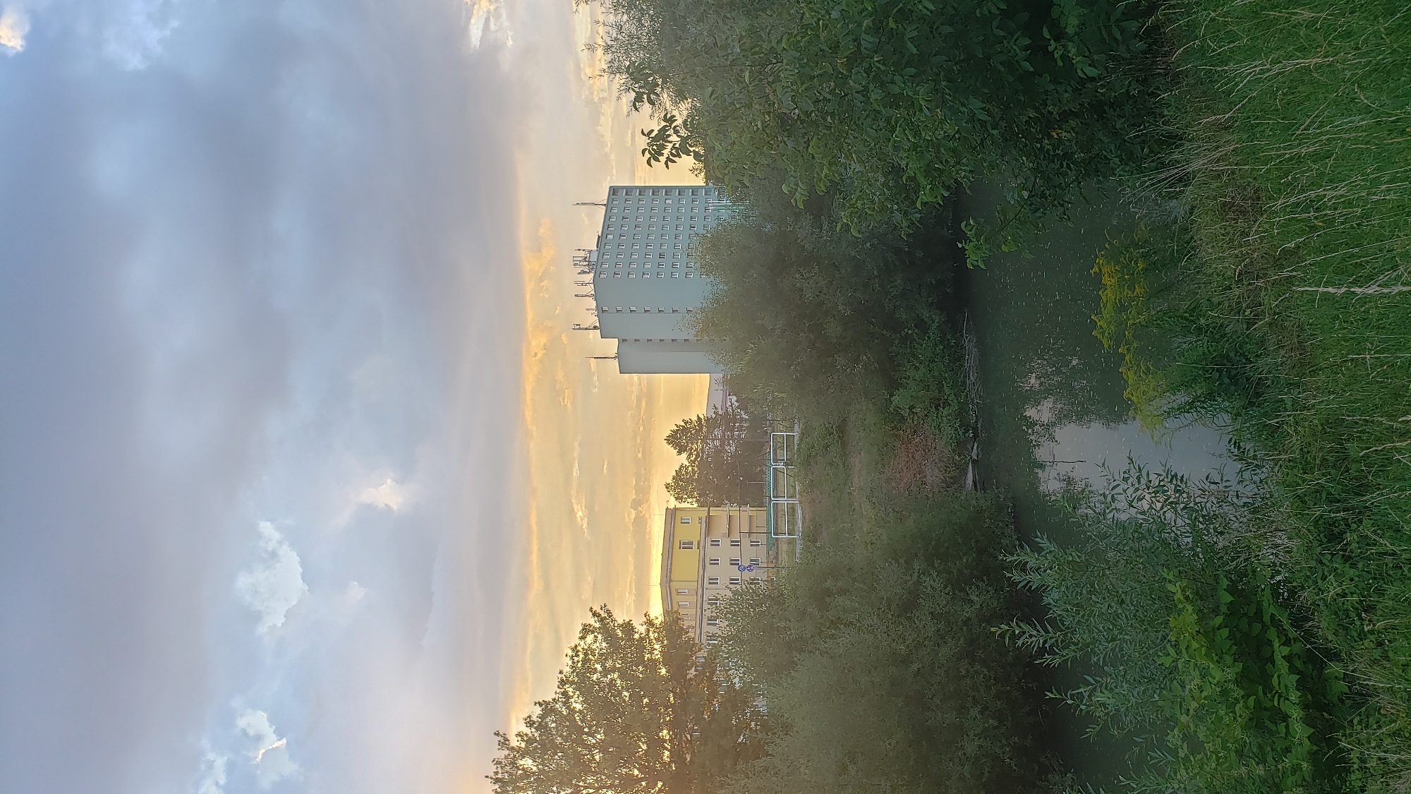 The image depicts an outdoor scene, likely at dusk, overlooking a body of water with dense green vegetation along its banks. In the background, there are two multi-story buildings. The building on the left is shorter and painted yellow with white accents. The building on the right is taller and light blue, with numerous windows and several antennas or communication towers on its roof. The sky is filled with dramatic clouds, with patches of warm light, possibly from the setting sun, breaking through. This scene is located in the city of Prešov, Slovakia, specifically near the Sekčov housing estate.