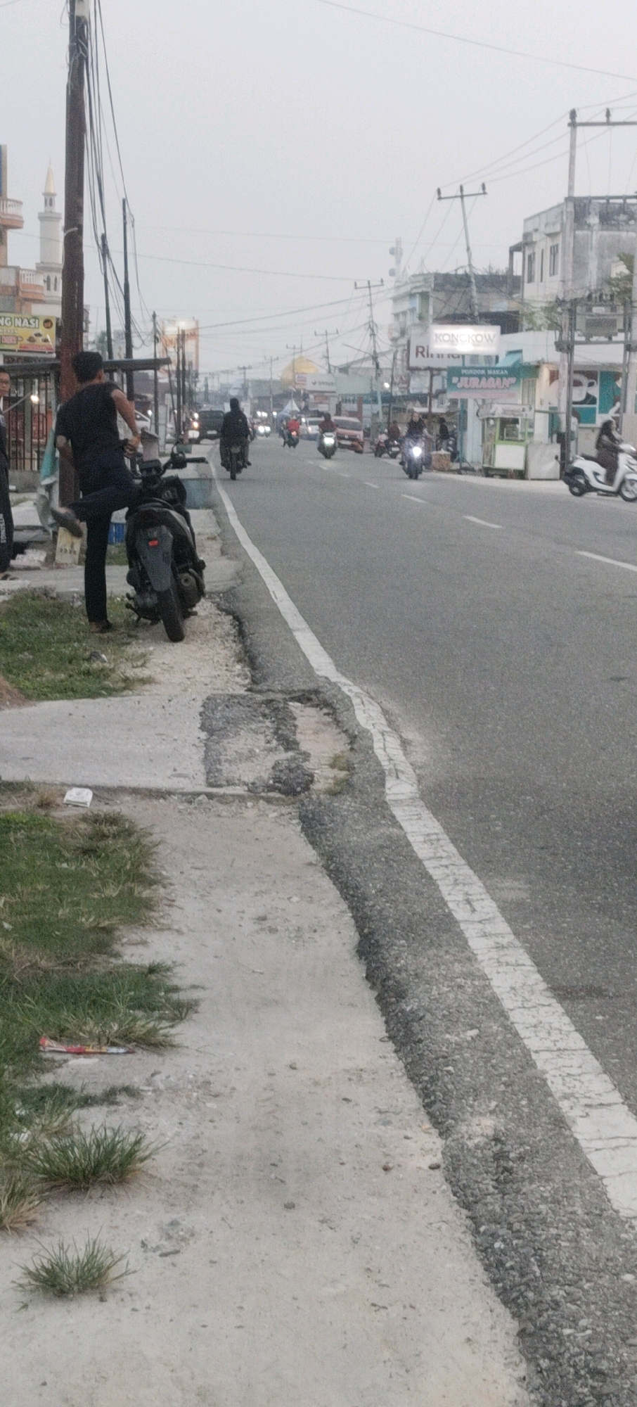 The image depicts a street scene in Sungai Pakning, Indonesia, under an overcast sky. A man in a black t-shirt and pants is standing next to a parked scooter on the side of a paved road. He is facing away from the camera and appears to be holding onto the scooter's handlebars with his right hand, with his left leg extended behind him. The scooter is dark-colored and positioned on a dirt and grass shoulder next to a concrete sidewalk.
The road is relatively wide and has a white dashed line marking the lane. Several other vehicles, primarily motorcycles, are visible on the road, with some appearing to be in motion. Buildings line the side of the road, displaying various signage. One sign reads "KONGKOW" and another indicates "PONDOK MAKAN JURAGAN". A minaret is visible in the distance on the left side of the image, suggesting the presence of a mosque. The overall environment appears to be a commercial or residential area with moderate traffic. The lighting suggests it is either early morning or late afternoon.