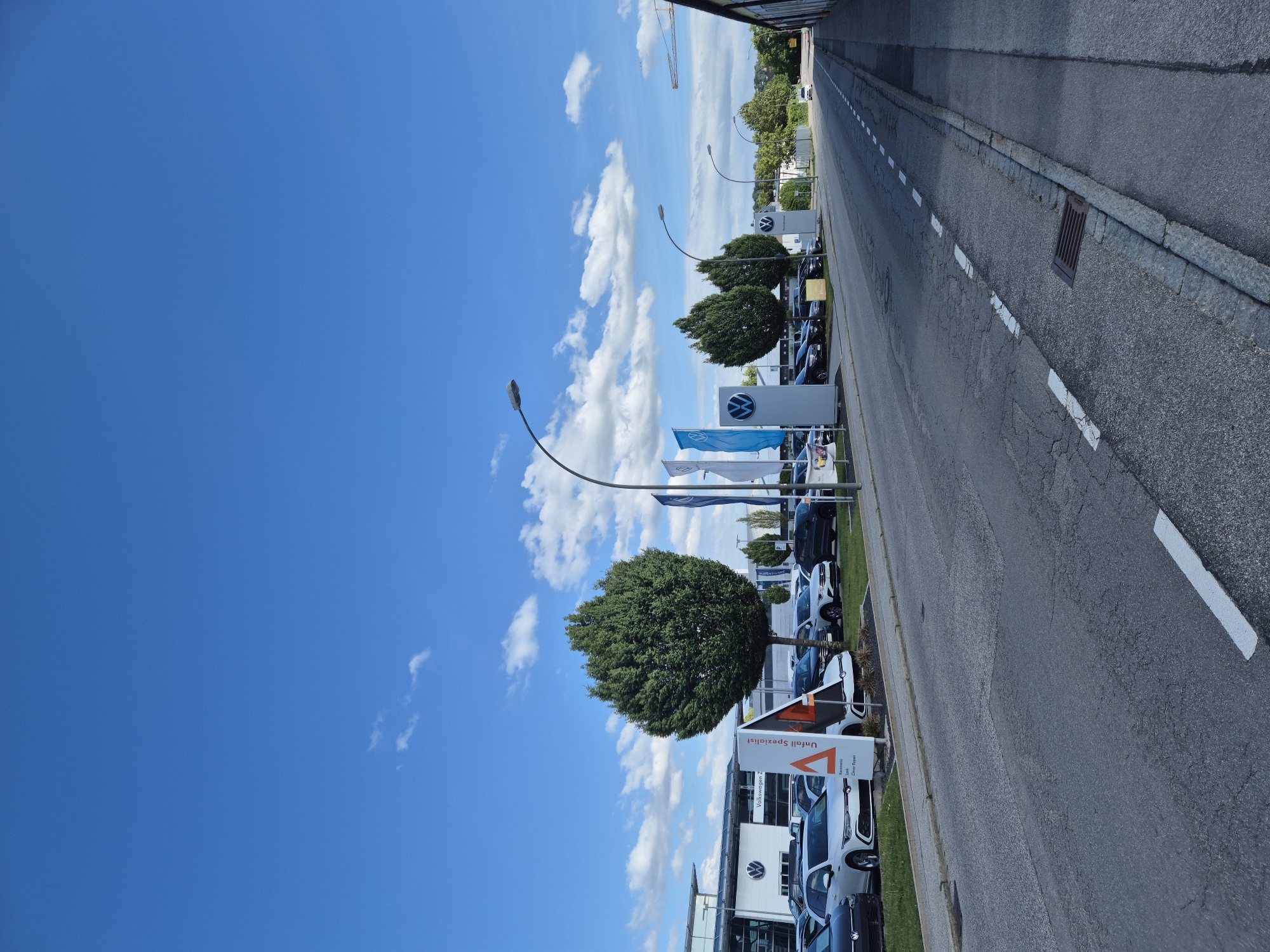 The image depicts a street scene in Landshut, Germany, likely in a commercial area given the presence of a Volkswagen dealership. The sky is a vibrant blue with scattered white clouds. In the foreground, a two-lane asphalt road runs from the lower left to the upper right of the frame. The road surface shows signs of wear and tear with visible cracks and patches. White dashed lines delineate the lanes. To the right of the road, a concrete sidewalk is visible, bordered by a stone curb. A metal grate is set into the road near the curb.
On the left side of the road, a Volkswagen dealership is apparent. Several Volkswagen cars are parked in rows on a landscaped area in front of the building, which features a modern architectural design with large windows. A prominent sign displays the Volkswagen logo, and another sign reads "Unfall Spezialist" (Accident Specialist) with additional text in smaller font indicating "Karosserie," "Lack," and "Clever Repair." Two tall, cone-shaped evergreen trees stand near the dealership. Blue and white flags, also featuring the Volkswagen logo, are displayed on poles. Streetlights are positioned along the road.
Further down the street, more trees and buildings are visible, suggesting a suburban or industrial edge of the city. In the distance, a yellow construction crane can be seen against the sky. The overall impression is a bright, sunny day at a car dealership on a city street. No people are visible in the image.